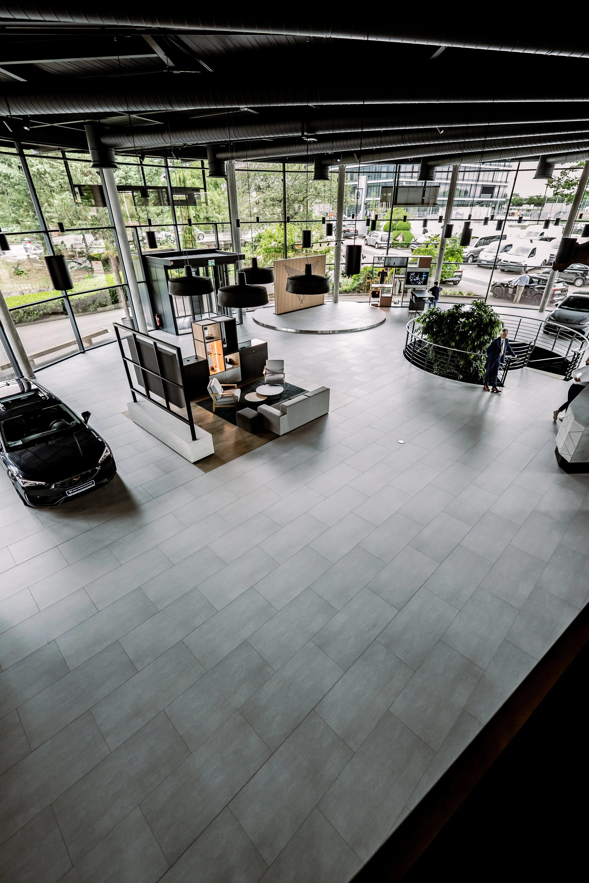 Große, moderne Autohändlerhalle mit Parkplätzen, Verkaufsraum und Eingangsbereich, Blick von oben, mit Autos, Möbeln und Personen im Innenraum.