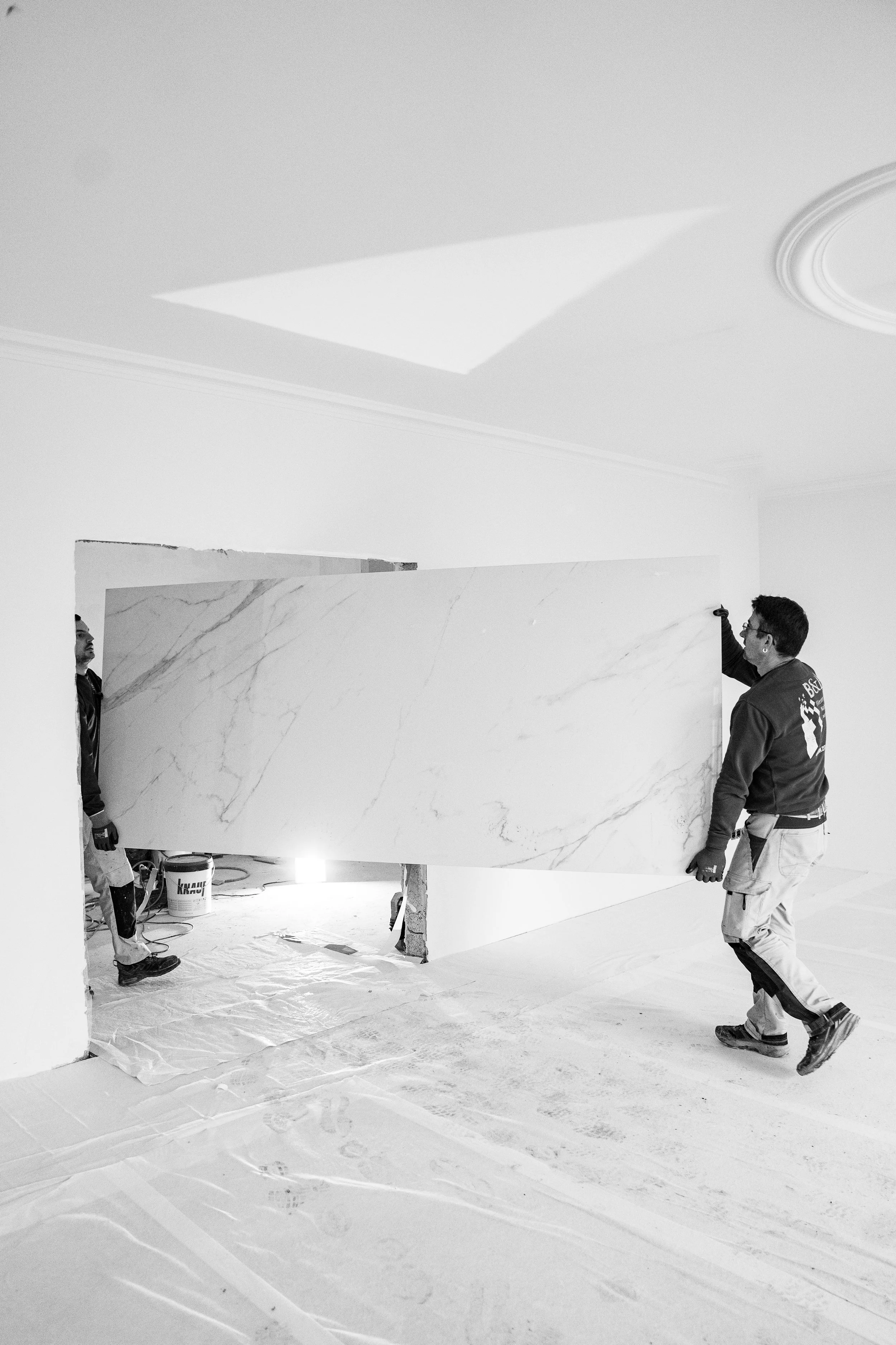 Zwei Arbeiter tragen eine große Wandplatte in einem renovierten Innenraum. Der Raum ist unvollständig und es werden Bauarbeiten durchgeführt.