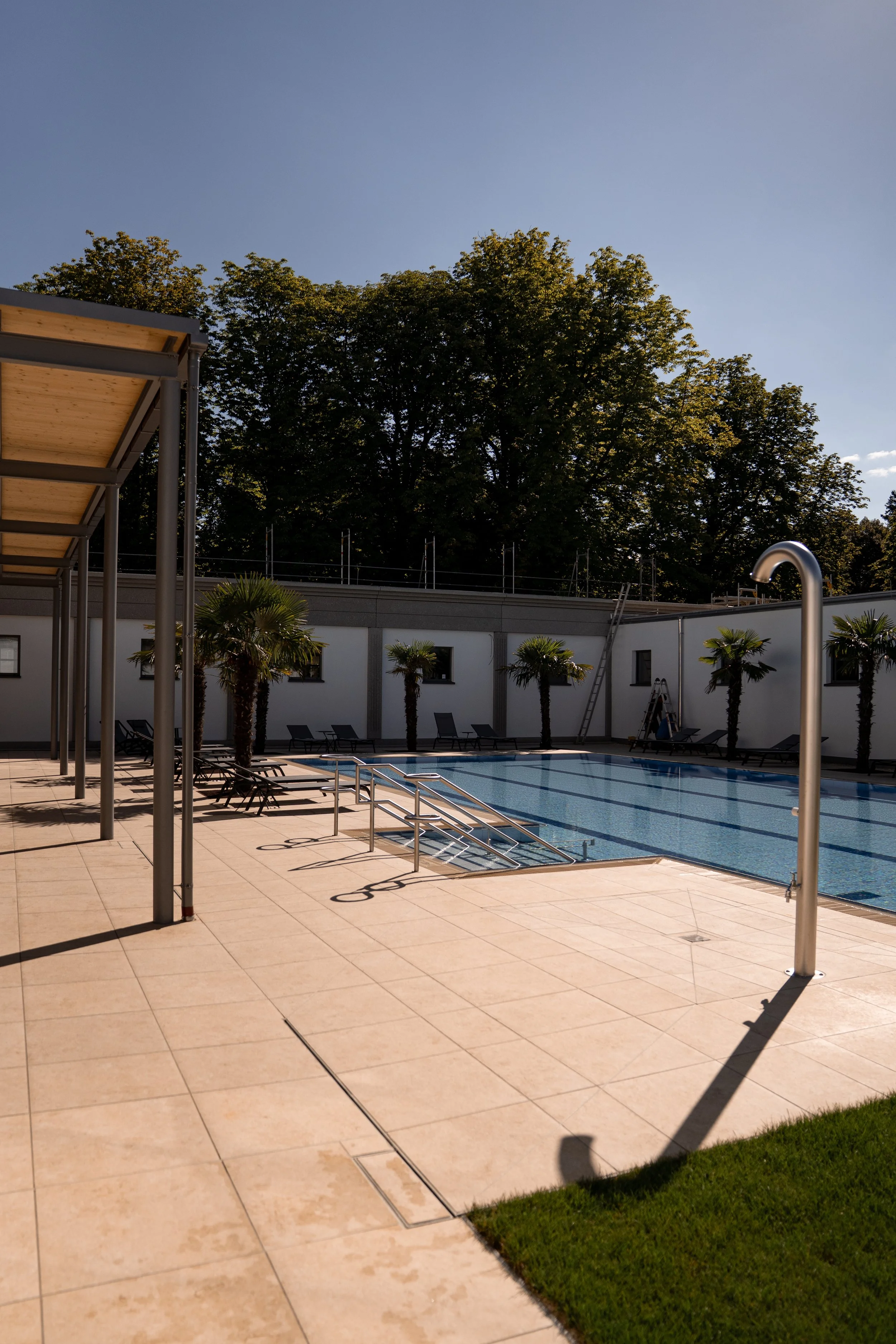 Ein Swimmingpool im Innenhof mit Liegestühlen, Palmen und Sonnenschirmen, umgeben von hohen Bäumen und einer weißen Mauer im Hintergrund.