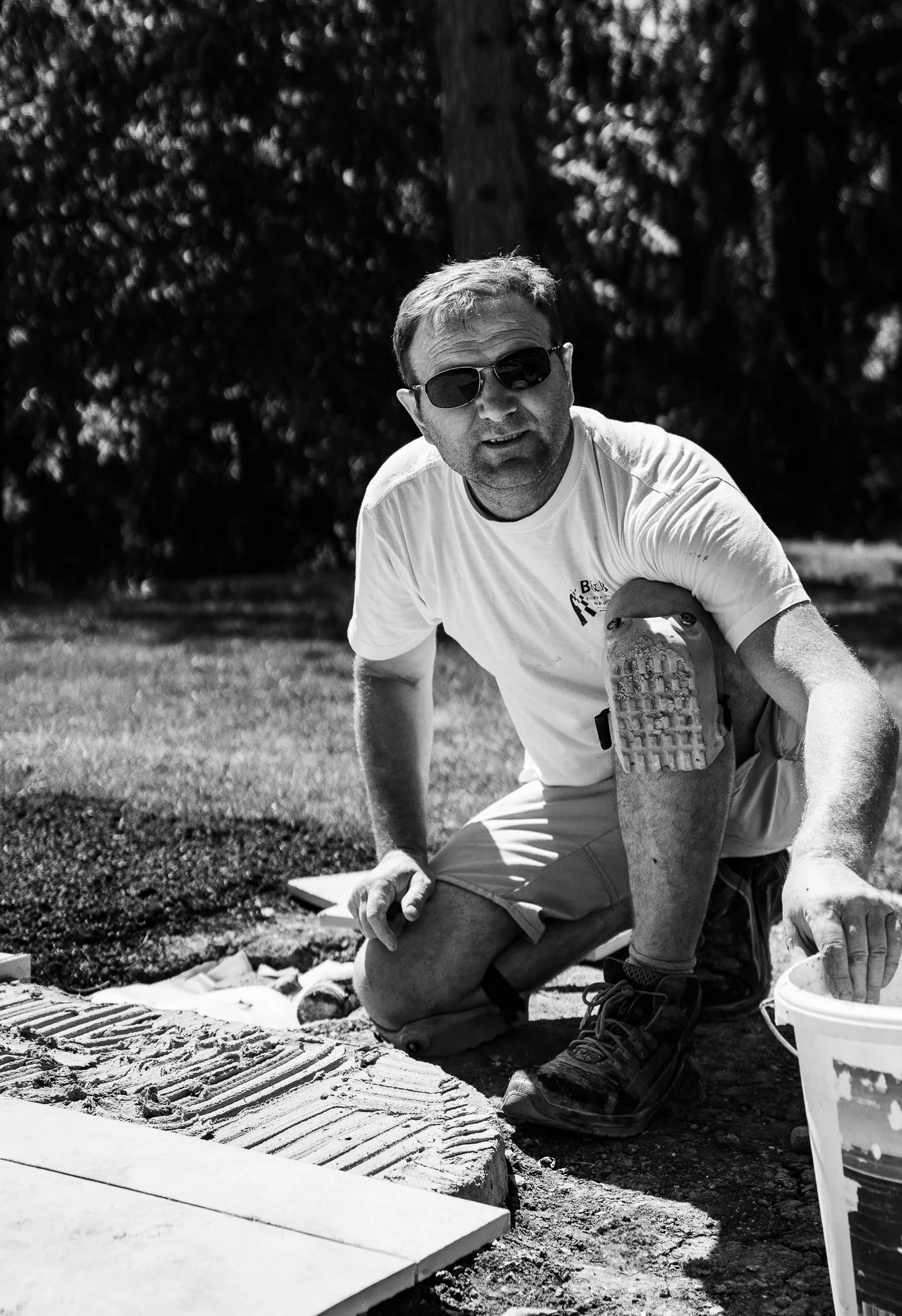 Mann beim Fliesenzuschnitt im Freien, trägt Sonnenbrille, weißes T-Shirt und Shorts, arbeitet an Terrassengestaltung.