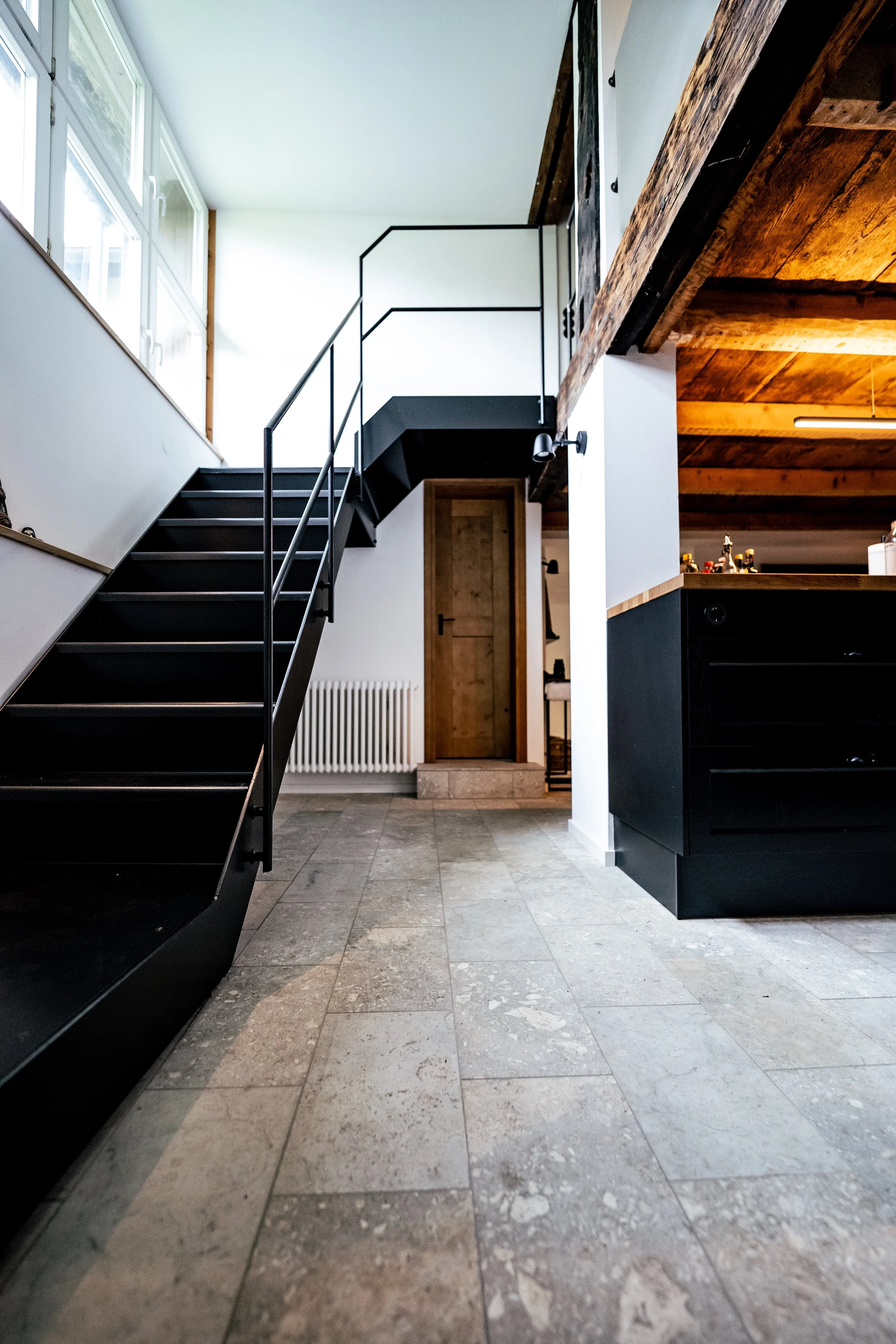 Innenansicht eines modernen Hauses mit schwarzer Metalltreppe, Holzbalken und Fliesenboden.