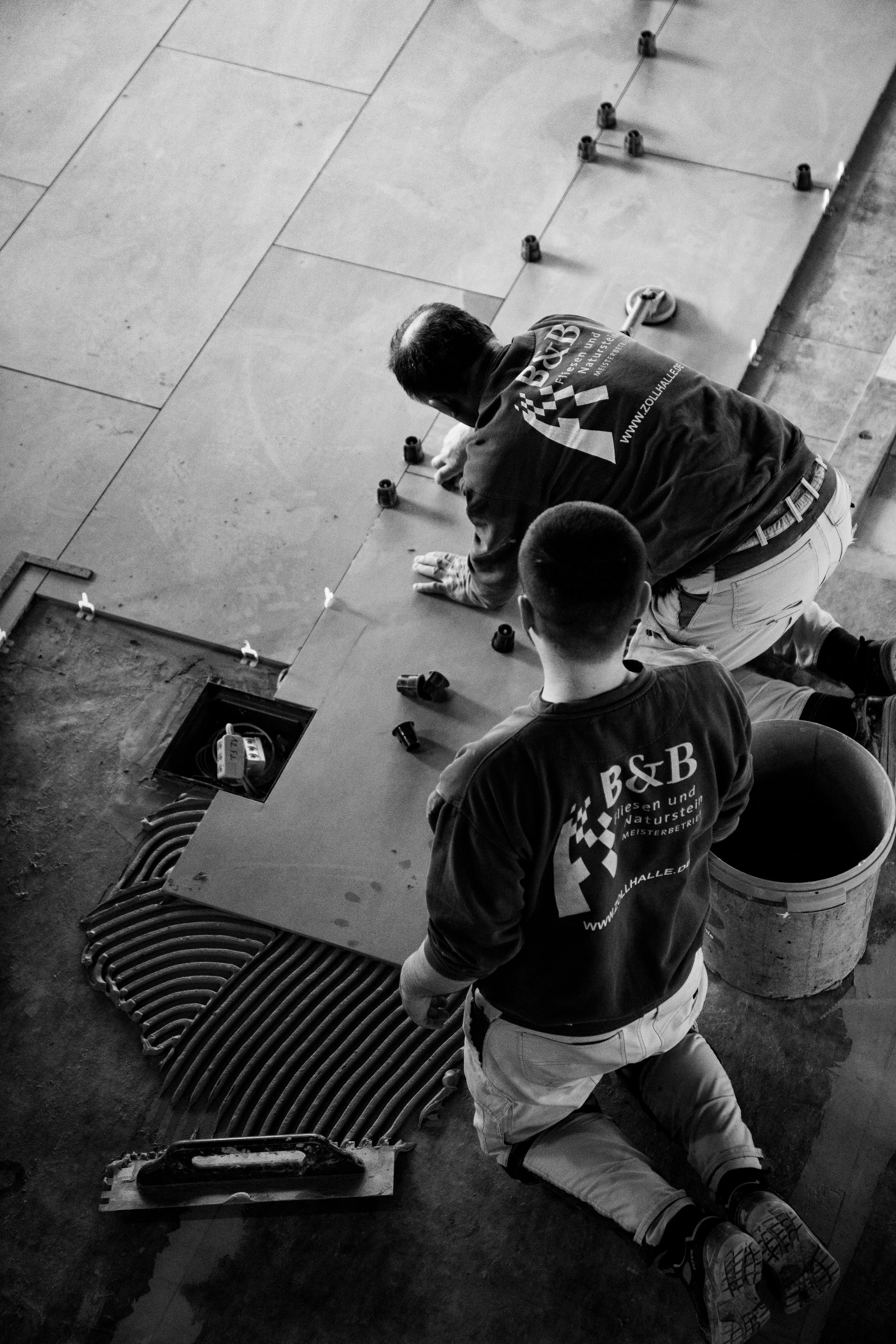 Zwei Arbeiter legen Fliesen auf den Boden, einer ist kniend, der andere ist in die Hocke gegangen, beide tragen T-Shirts mit dem Logo und der Webseite www.zollhalle.de.