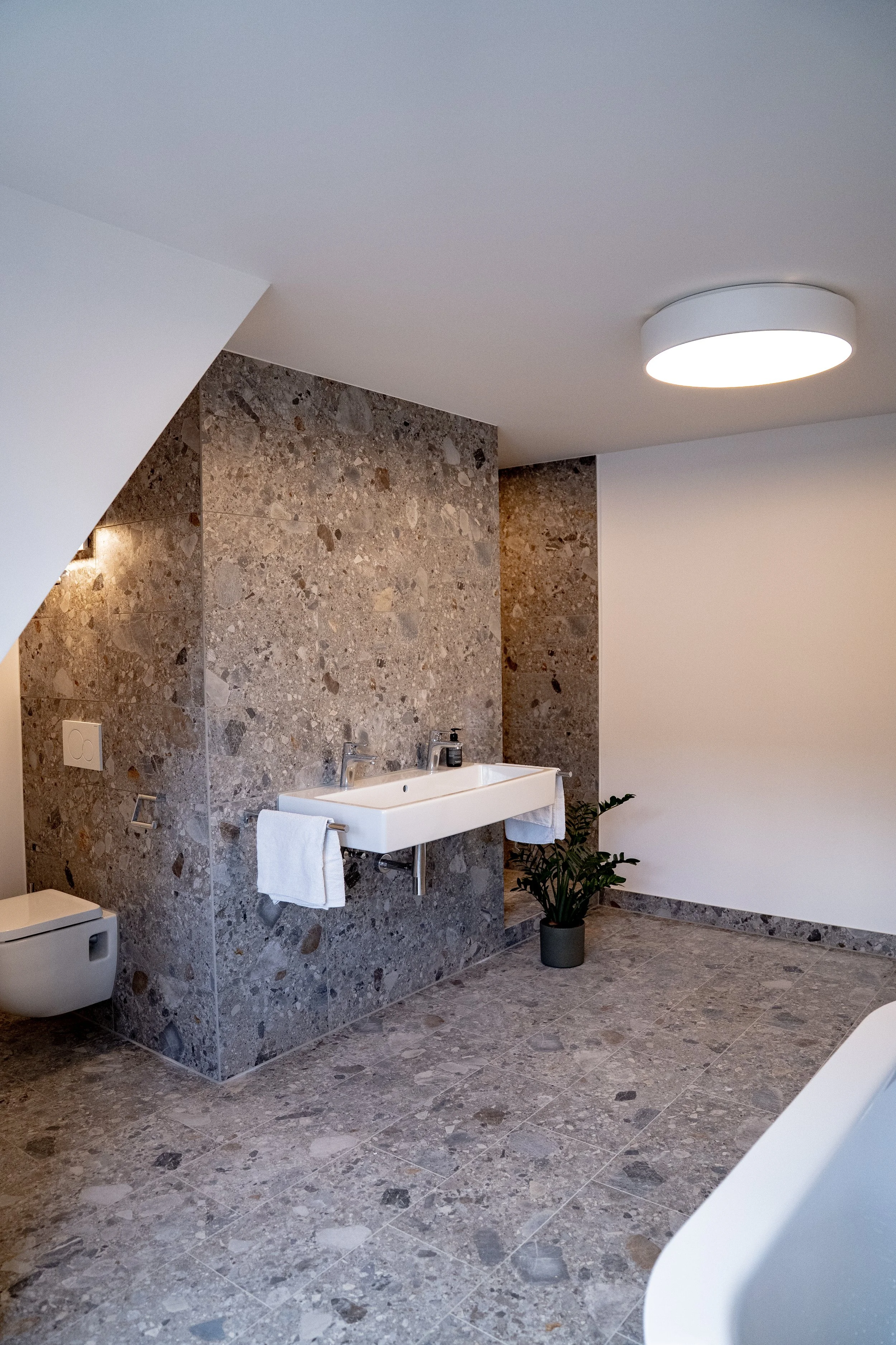 Modernes Badezimmer mit Waschbecken, Toilette und Steinfliesen, Pflanze und Deckenleuchte.