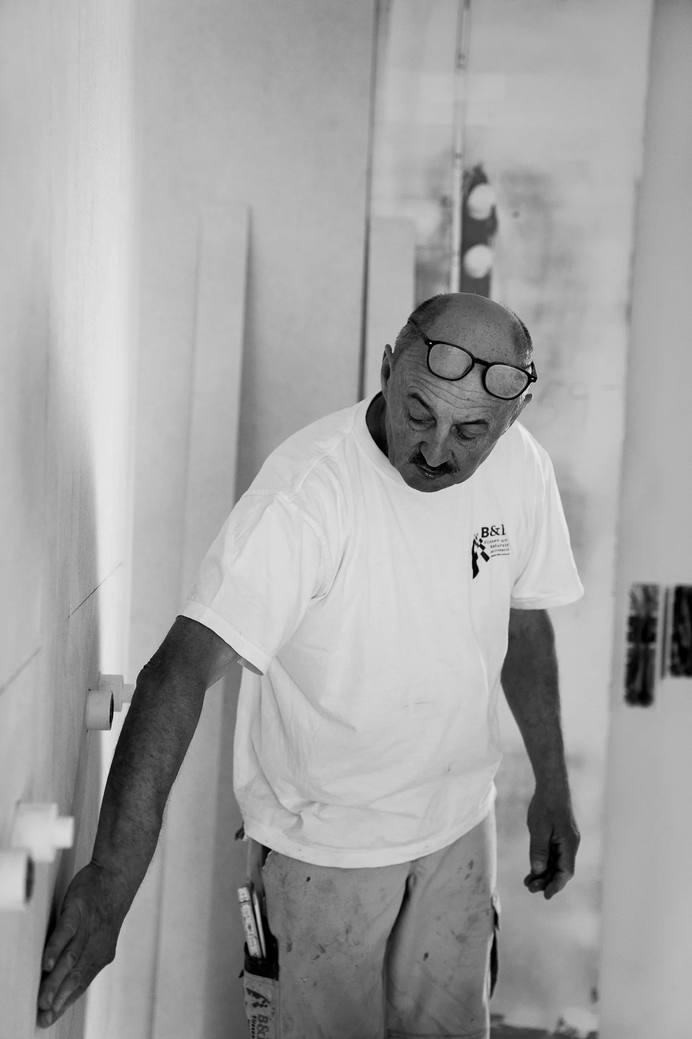 Älterer Mann mit Brille auf dem Kopf, trägt T-Shirt und Arbeitshose, arbeitet in einem Raum, vermutlich bei Renovierungsarbeiten.