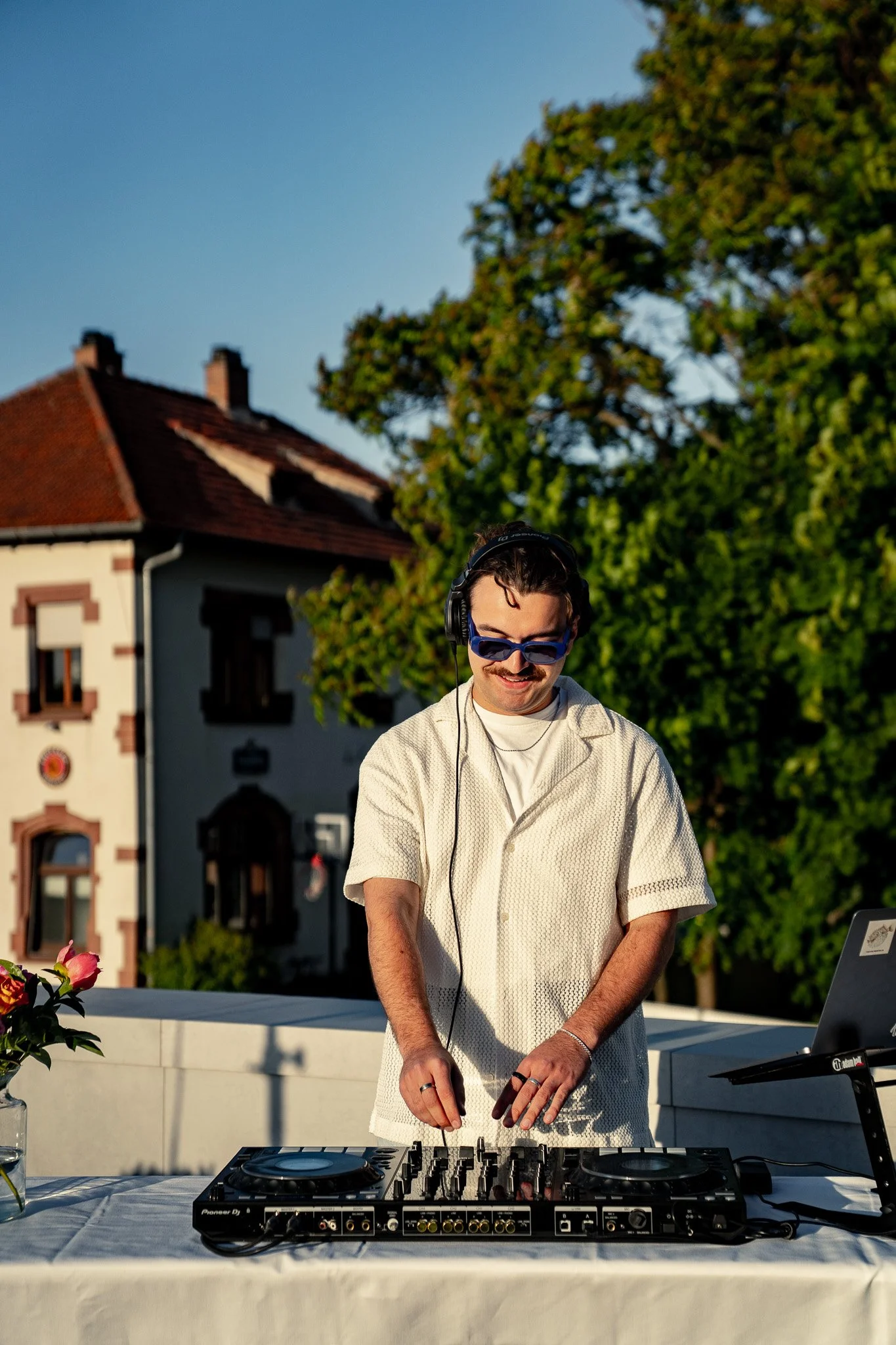 Ein Mann mit dunklen Haaren und Sonnenbrille, der an einem DJ-Controller auf einem weißen Tisch im Freien steckt, trägt einen weißen, kurzärmligen knopfblusenartigen Pullover und hört Musik mit Kopfhörern, während er lächelt.
