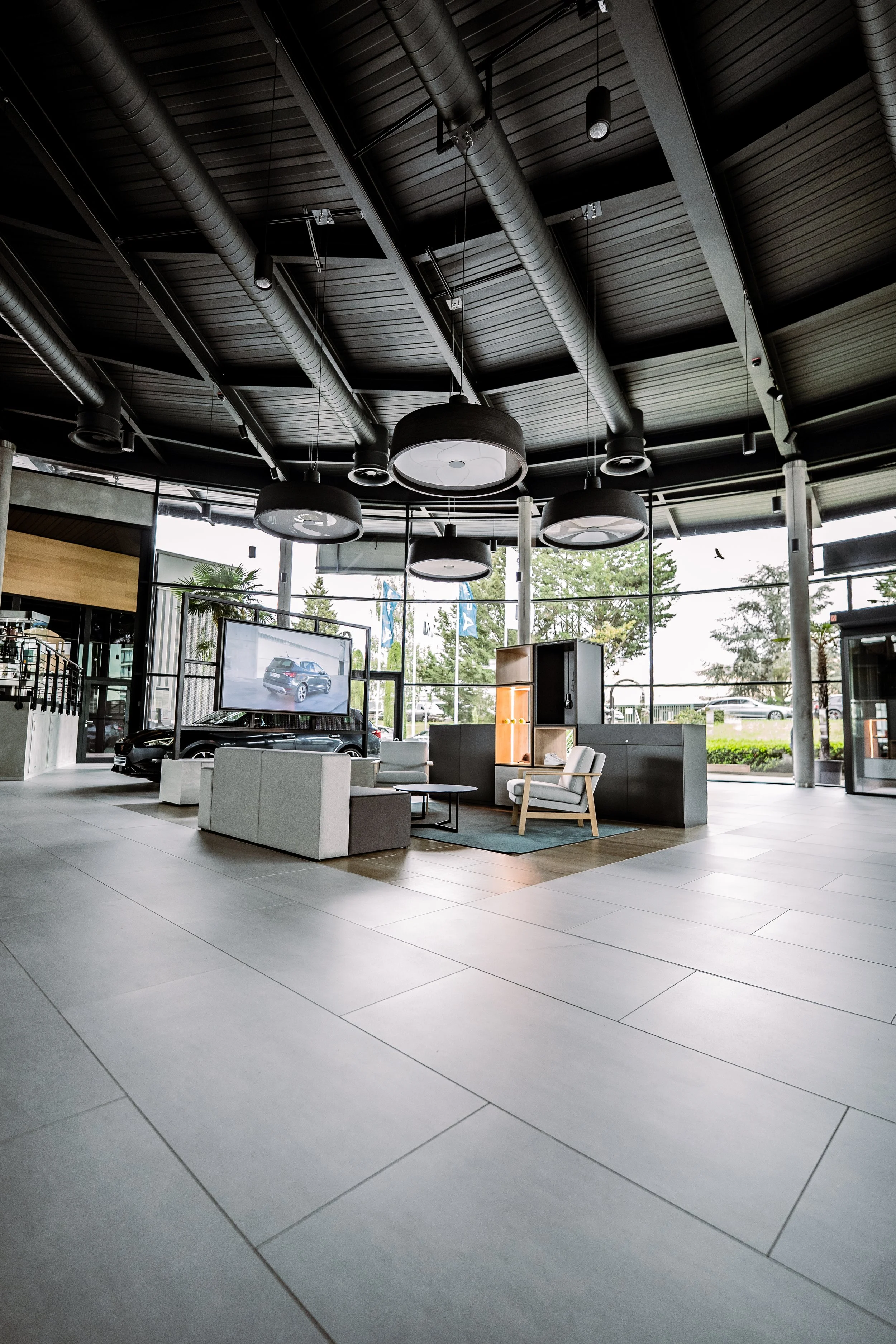 Moderne Autohandels Showroom mit Sitzbereich, Fernseher und großen Fenstern, durch die man Fahrzeuge im Freien sehen kann.