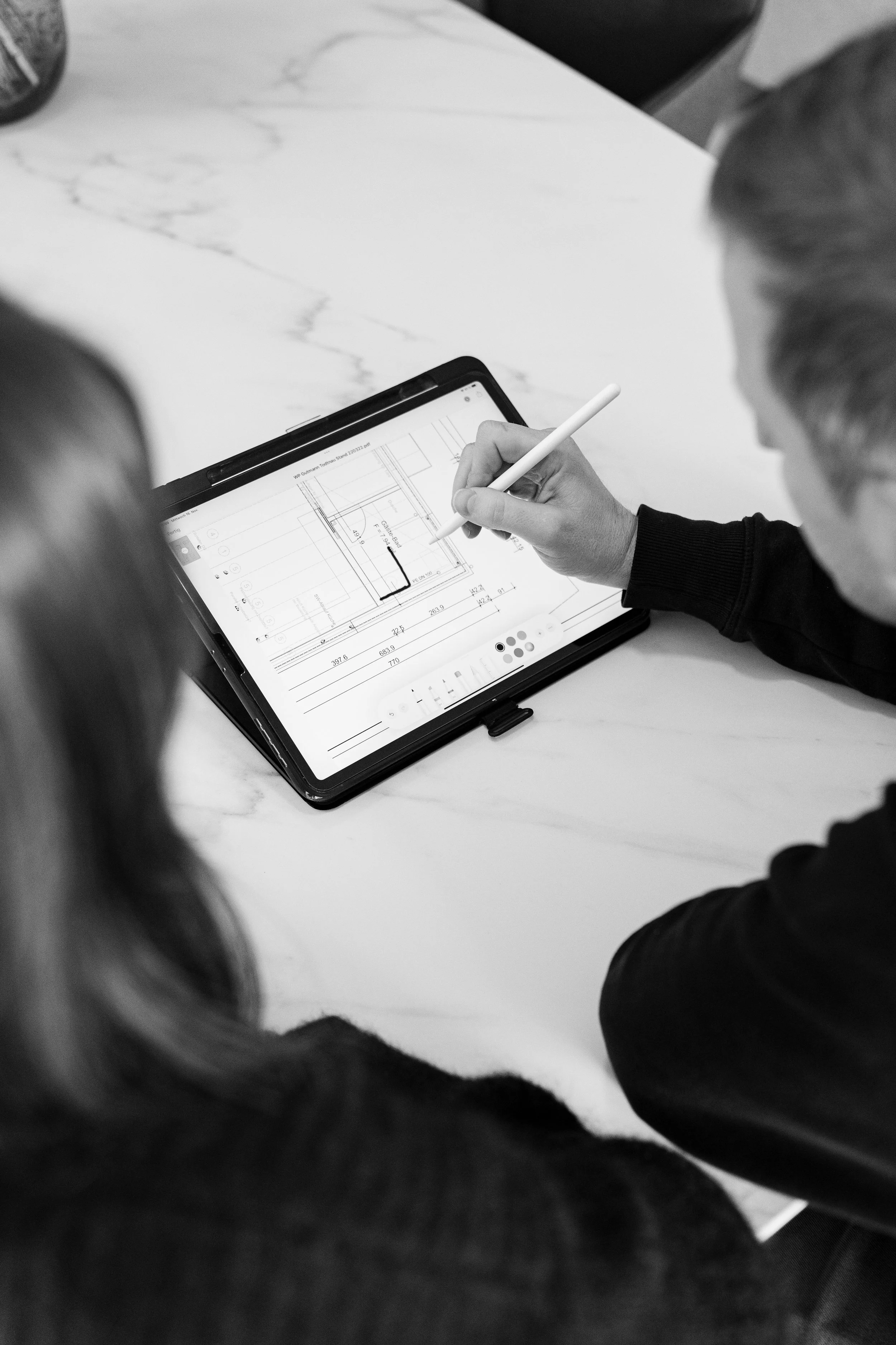 Zwei Personen betrachten einen digitalen Bauplan auf einem Tablet, wobei eine Person mit einem Stift auf den Bildschirm zeigt.