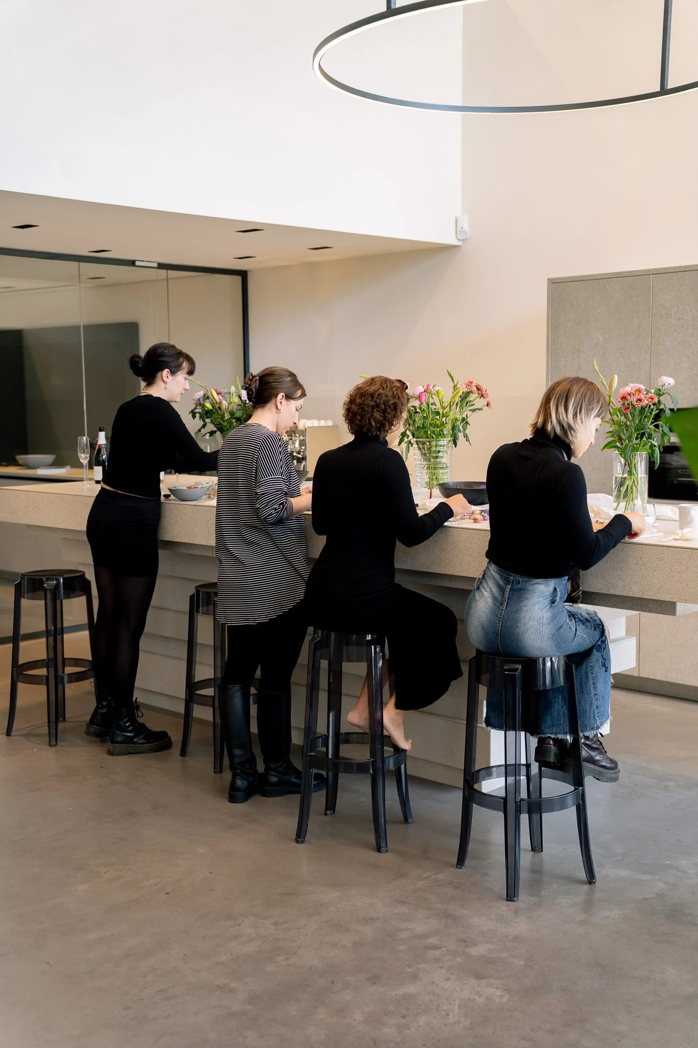 Vier Frauen sitzen an einer Theke im modernen, minimalistischen Raum, trinken Getränke und haben Blumen in Vasen vor sich.