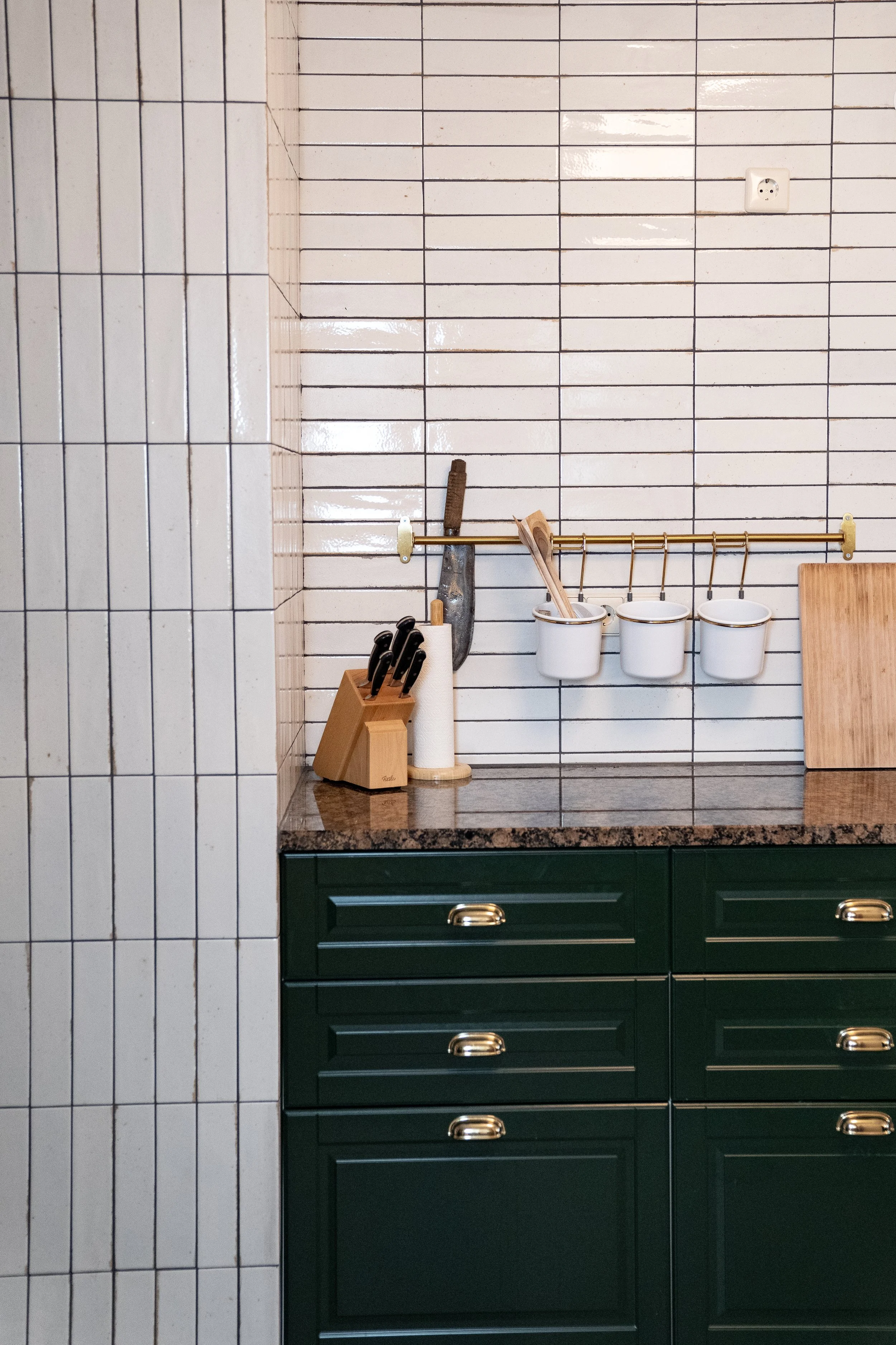 Küchenschrank mit Messerblock, Küchenrolle, Küchenutensilien, weißen Haken mit Tassen, Arbeitsplatte, Wandfliesen und Steckdose.