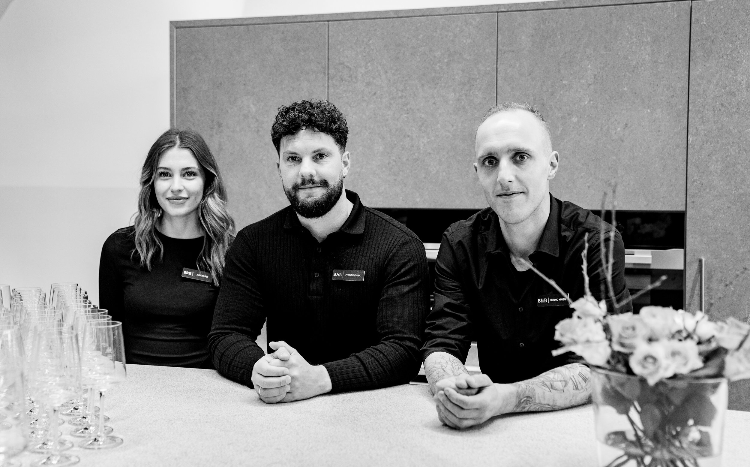 Drei Personen sitzen an einem Tisch, links eine Frau und in der Mitte und rechts zwei Männer, die alle schwarze Kleidung tragen.