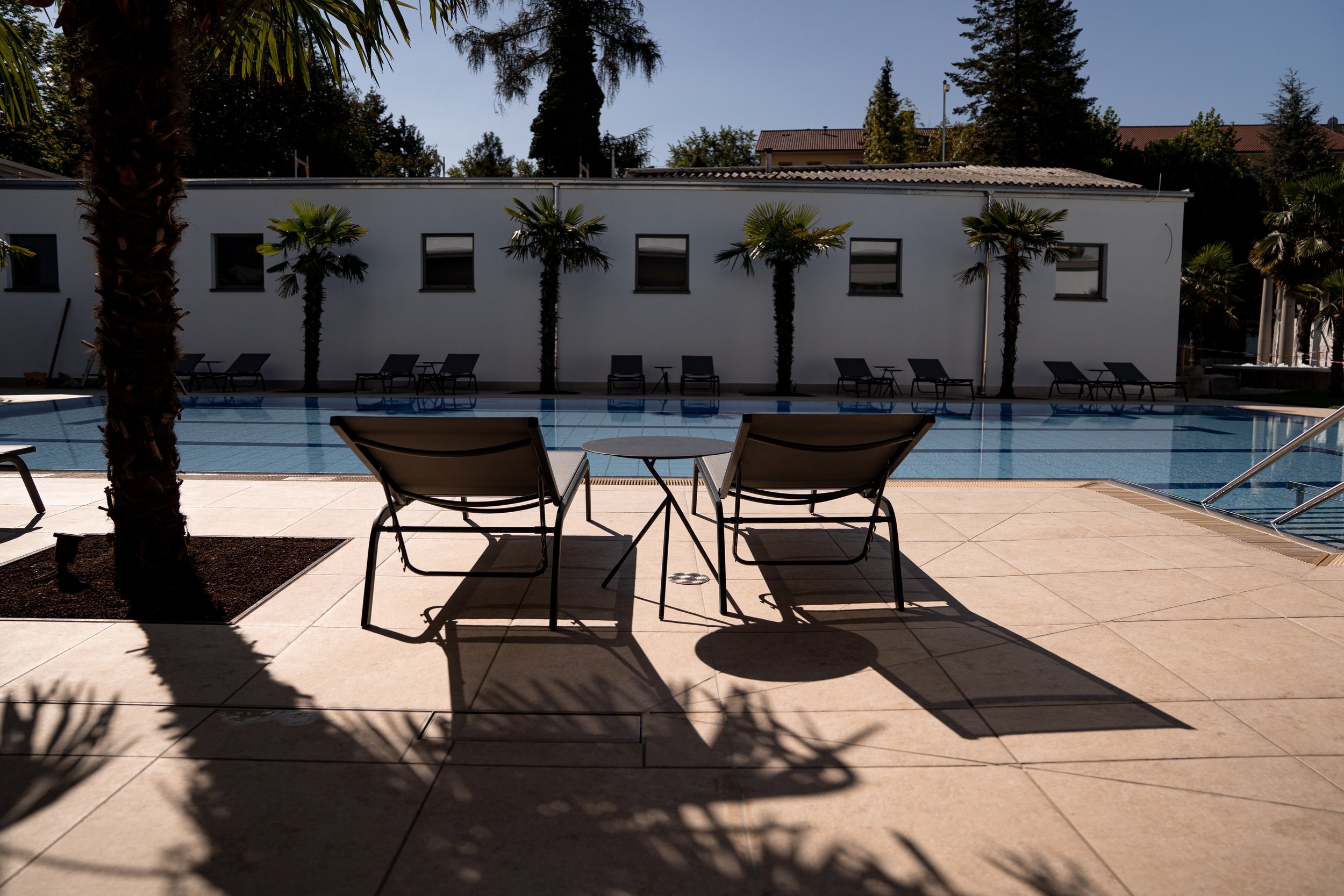 Ein leeres Schwimmbad im Freien mit Sonnenliegen und kleinen Tischen, umgeben von Palmen, in einem Hotelgarten bei Sonnenlicht.