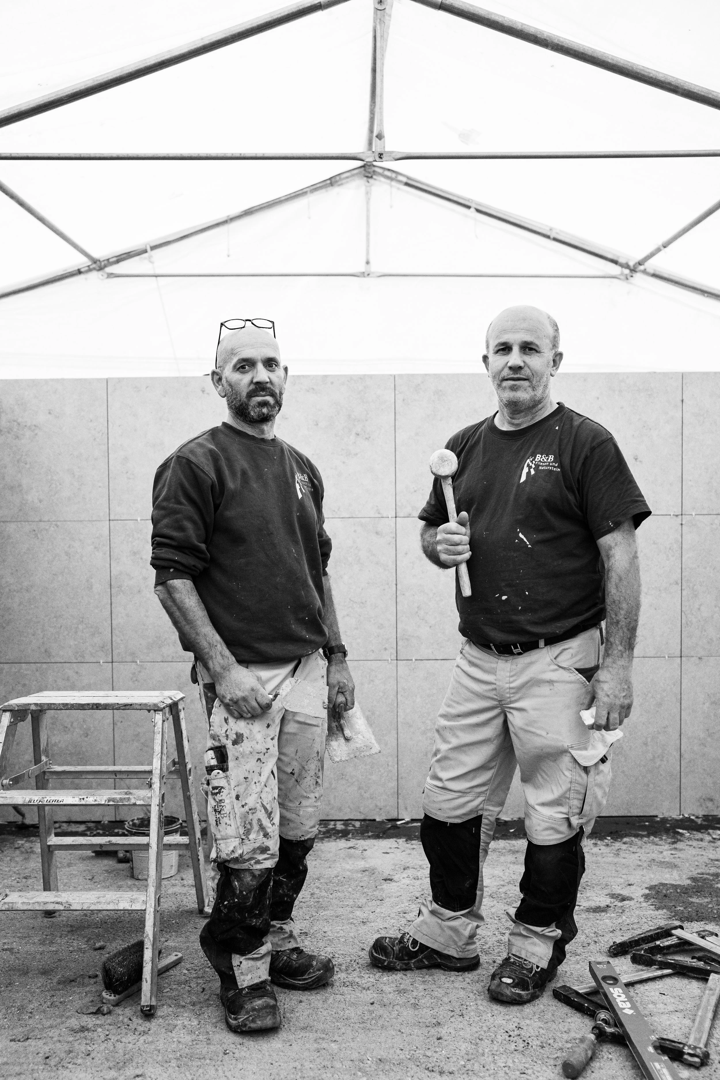 Zwei Bauarbeiter stehen in einer Baustelle, einer hält einen Hammer, beide tragen dunkle T-Shirts, die Bauarbeiterkleidung ist schmutzig, im Hintergrund sind Wandpaneele und ein Gerüst zu sehen.