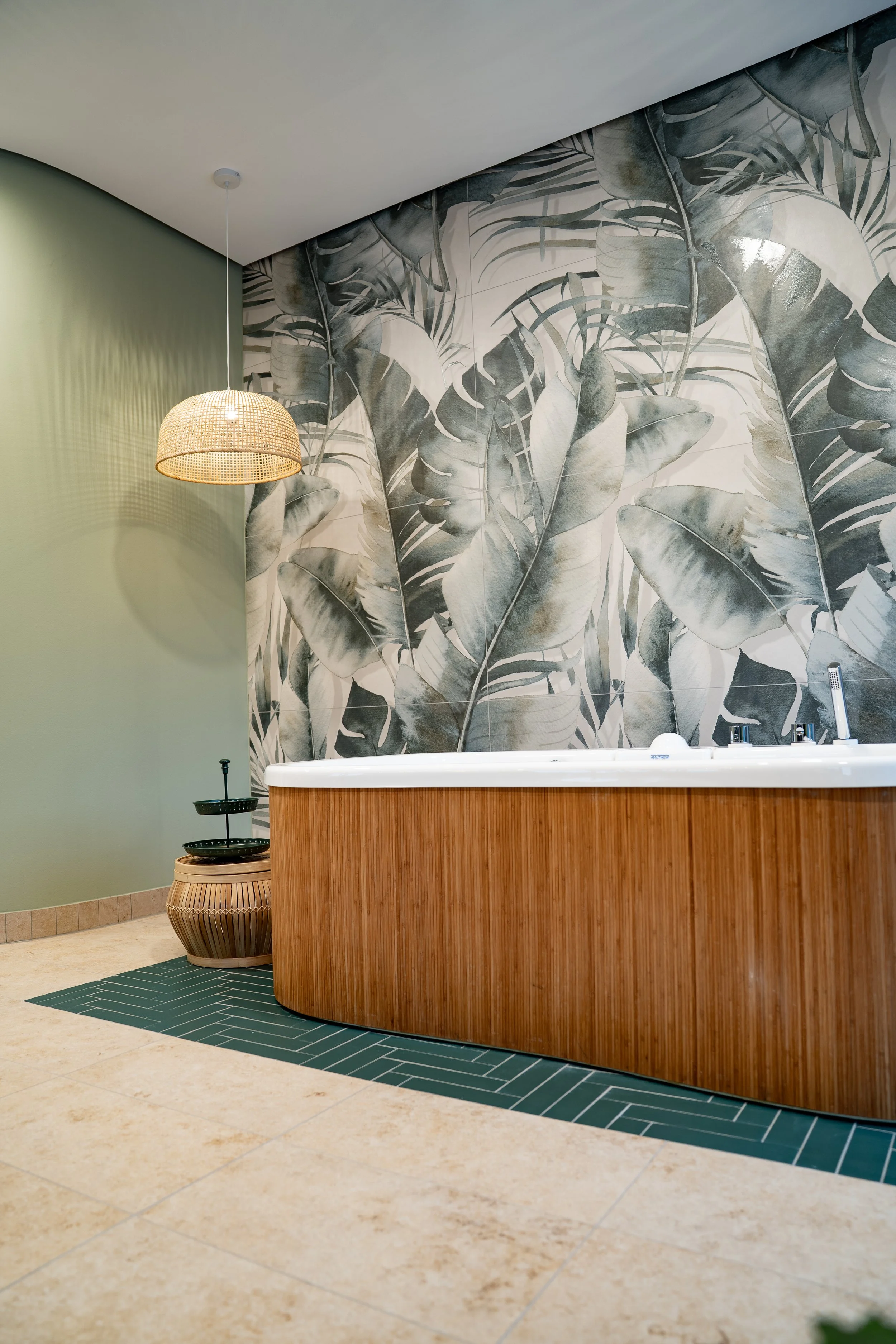 Ein Badezimmer mit einer Wanddekoration mit großen, schwarzen und weißen Blättern, einer runden Holzwandbadewanne, einem grünen Bodenbelag und einer Stehlampe mit Rattan-Schirm.