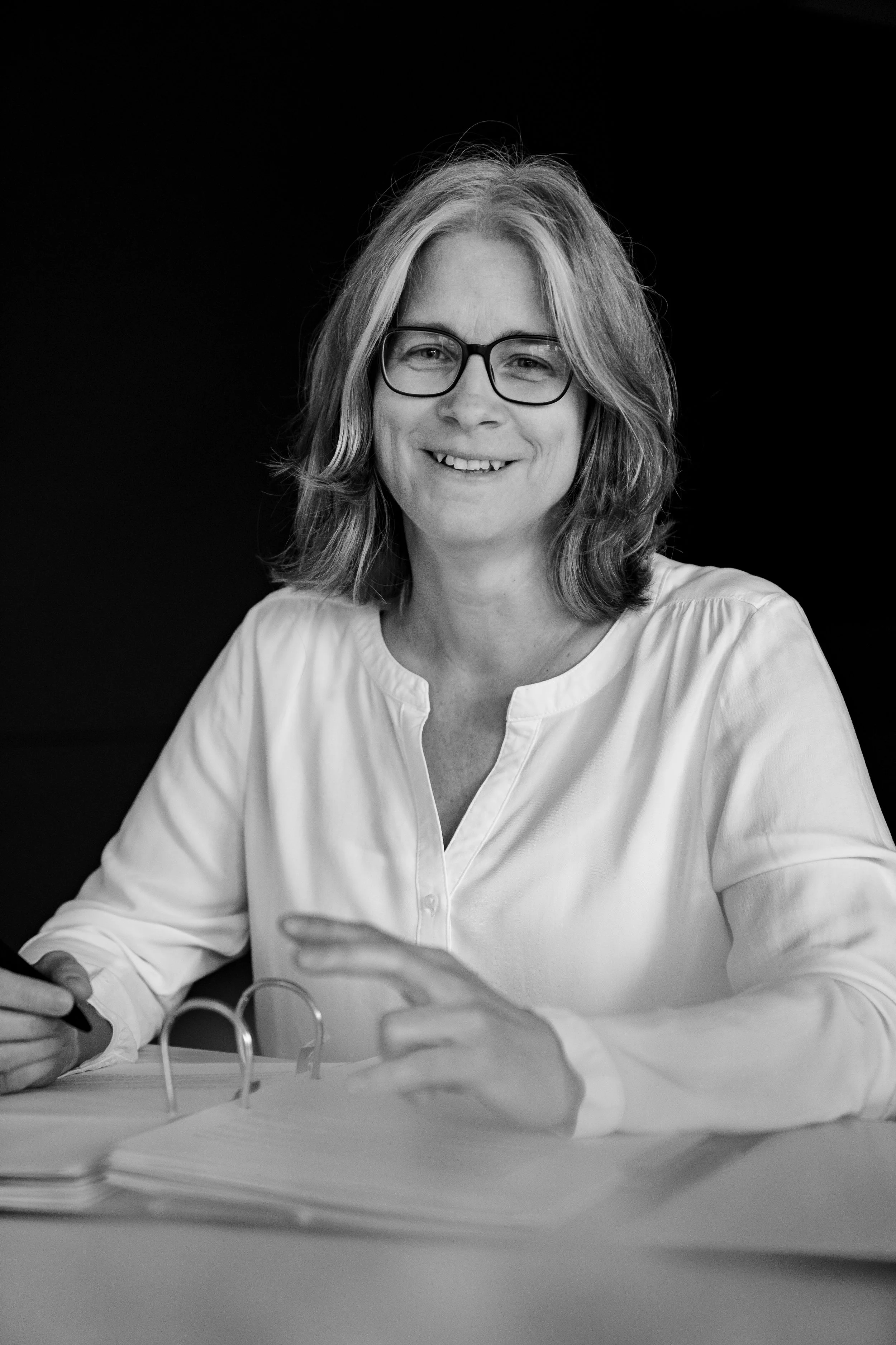 Schwarz-weiß-Porträt einer lächelnden Frau mit Brille, sitzend an einem Tisch mit Akten, gegen schwarzen Hintergrund.