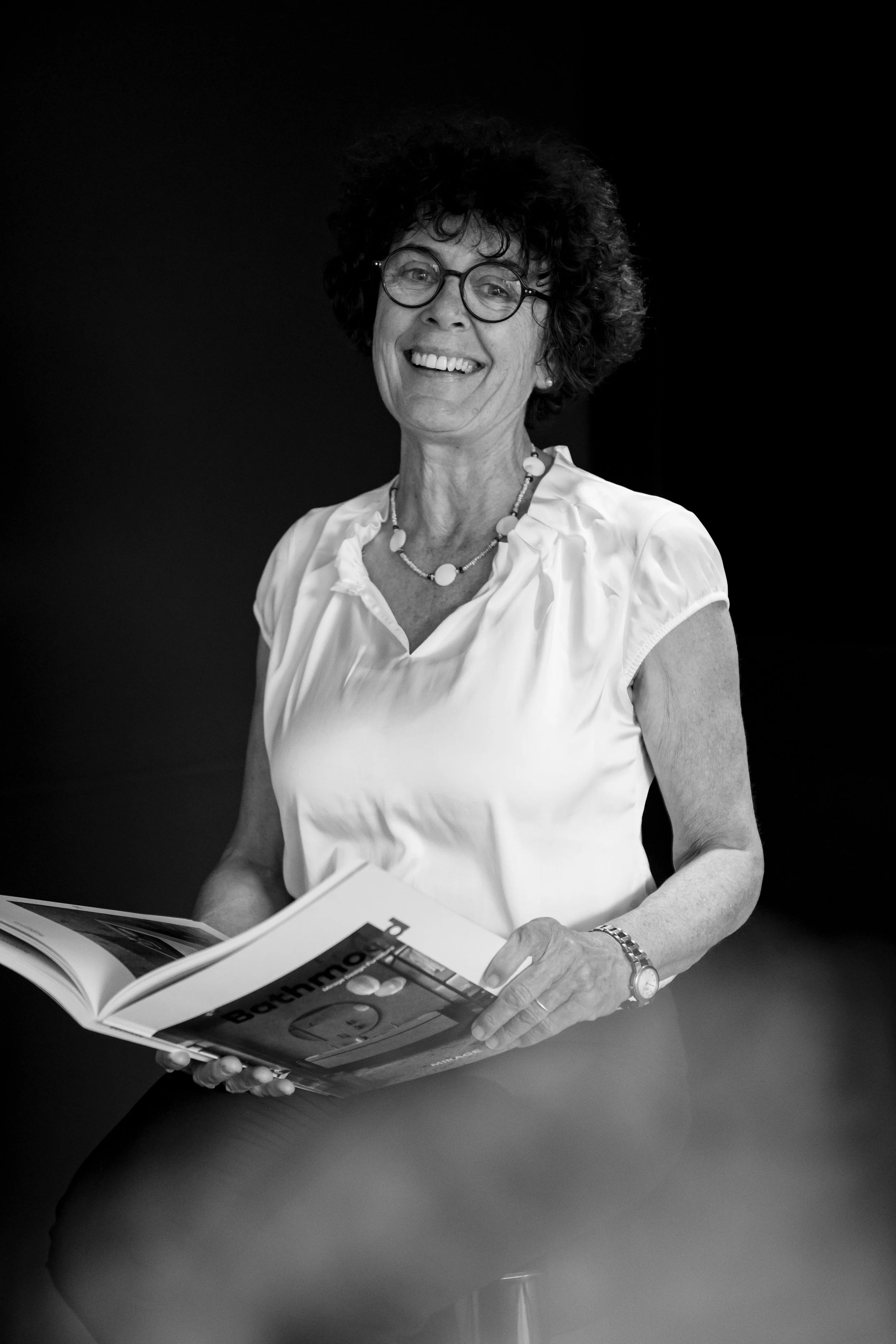 Eine lachende, ältere Frau mit Brille, kurzer dunkler Lockenfrisur, weißer Bluse, Halskette und Armband, die in einem Magazin blättert.