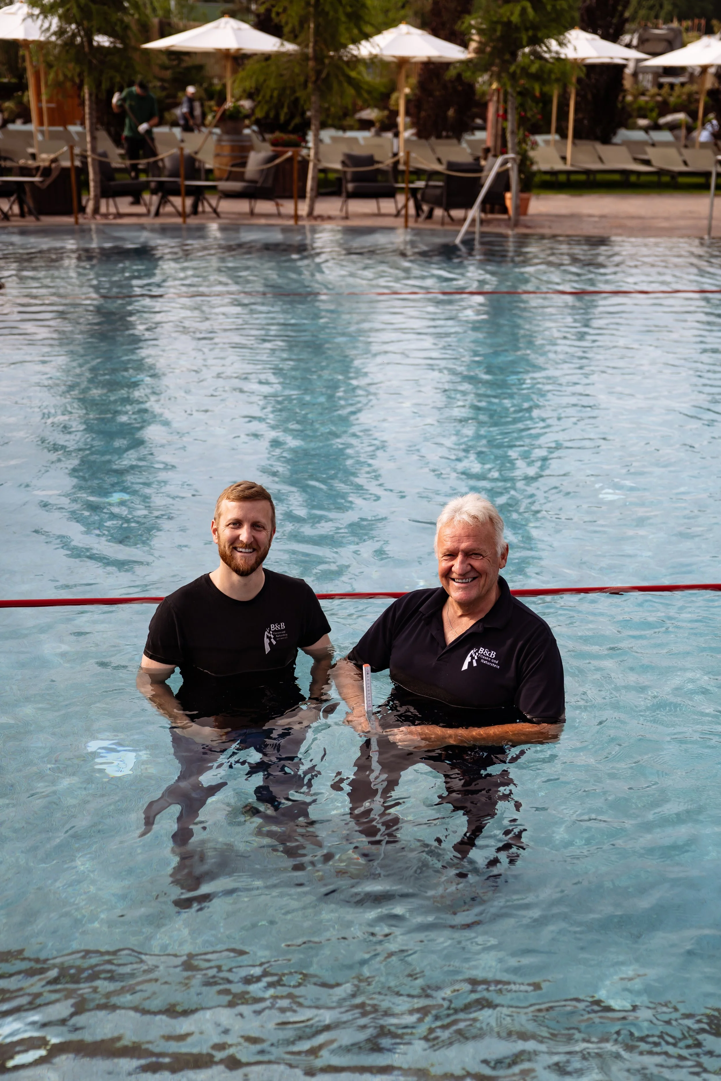Zwei Männer stehen im Pool, lächeln und tragen schwarze T-Shirts, im Hintergrund Sonnenschirme und Liegen.