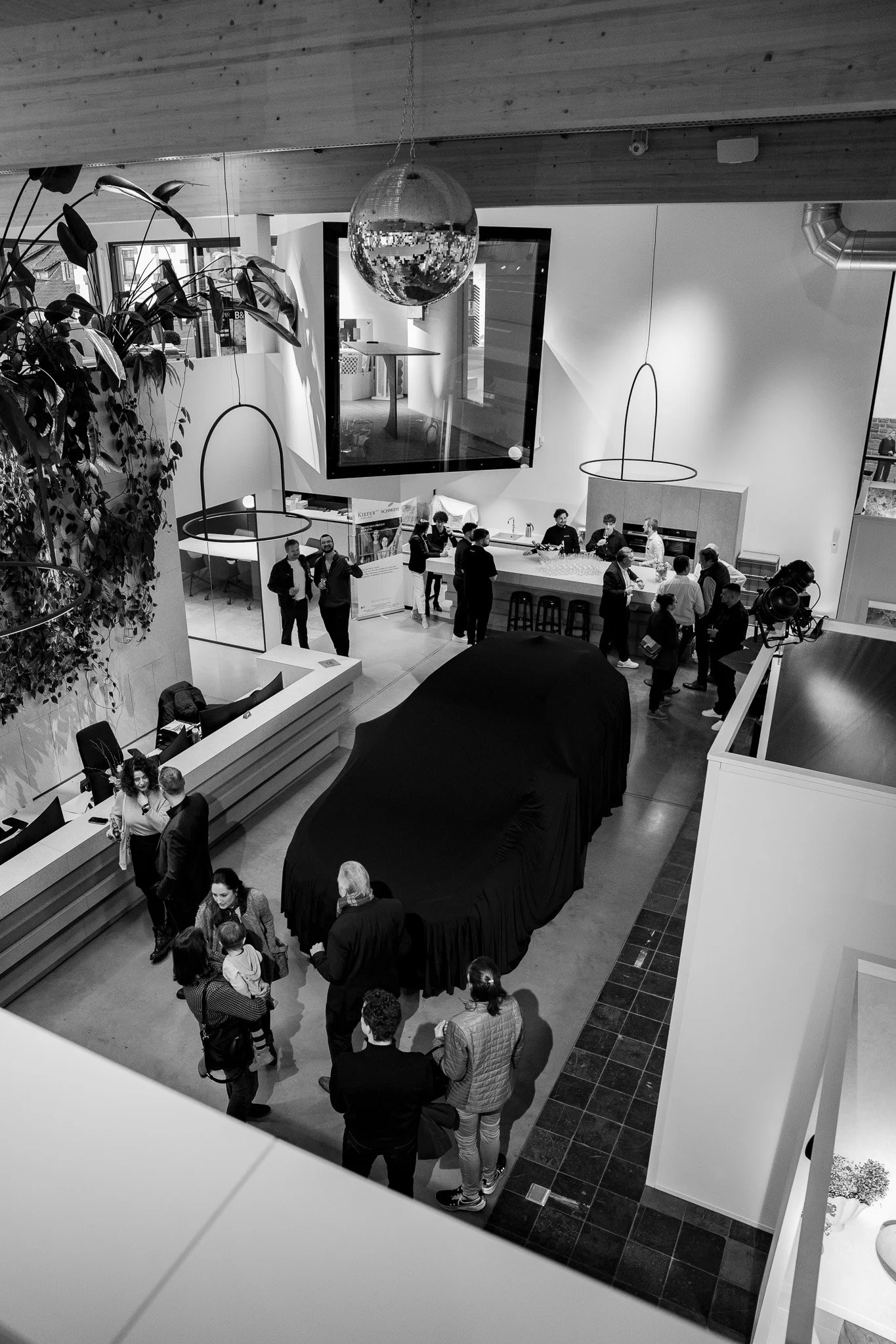 Veranstaltung in einem modernen, zweistöckigen Innenraum mit Menschen, die um ein abgedecktes Fahrzeug herumstehen, darunter auch Kinder, und einer Bar im Hintergrund.