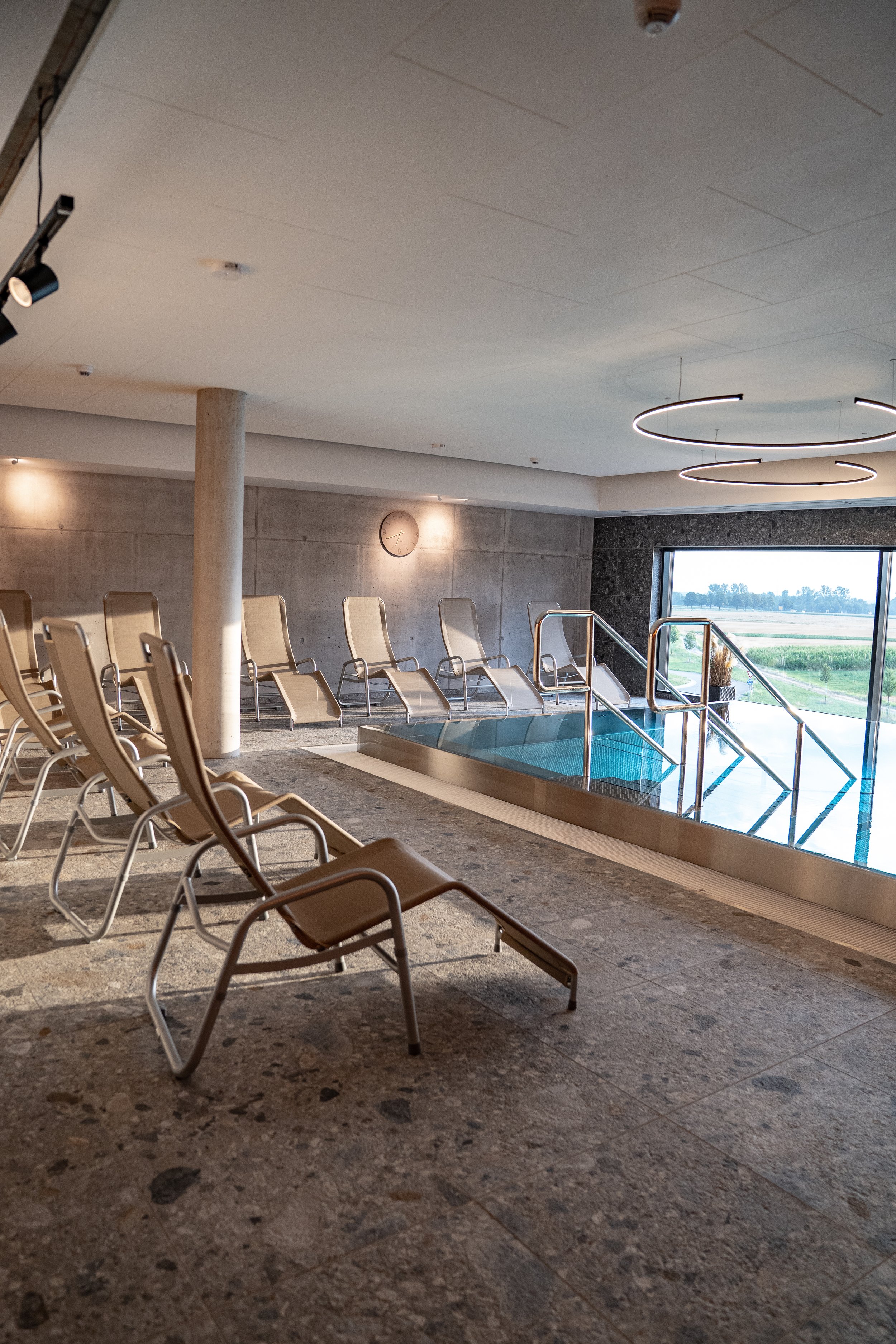 Ein Innenraum eines Spa- oder Wellnessbereichs mit Liegeflächen, einem kleinen Pool, großen Fenstern mit Blick ins Freie und moderner Beleuchtung.