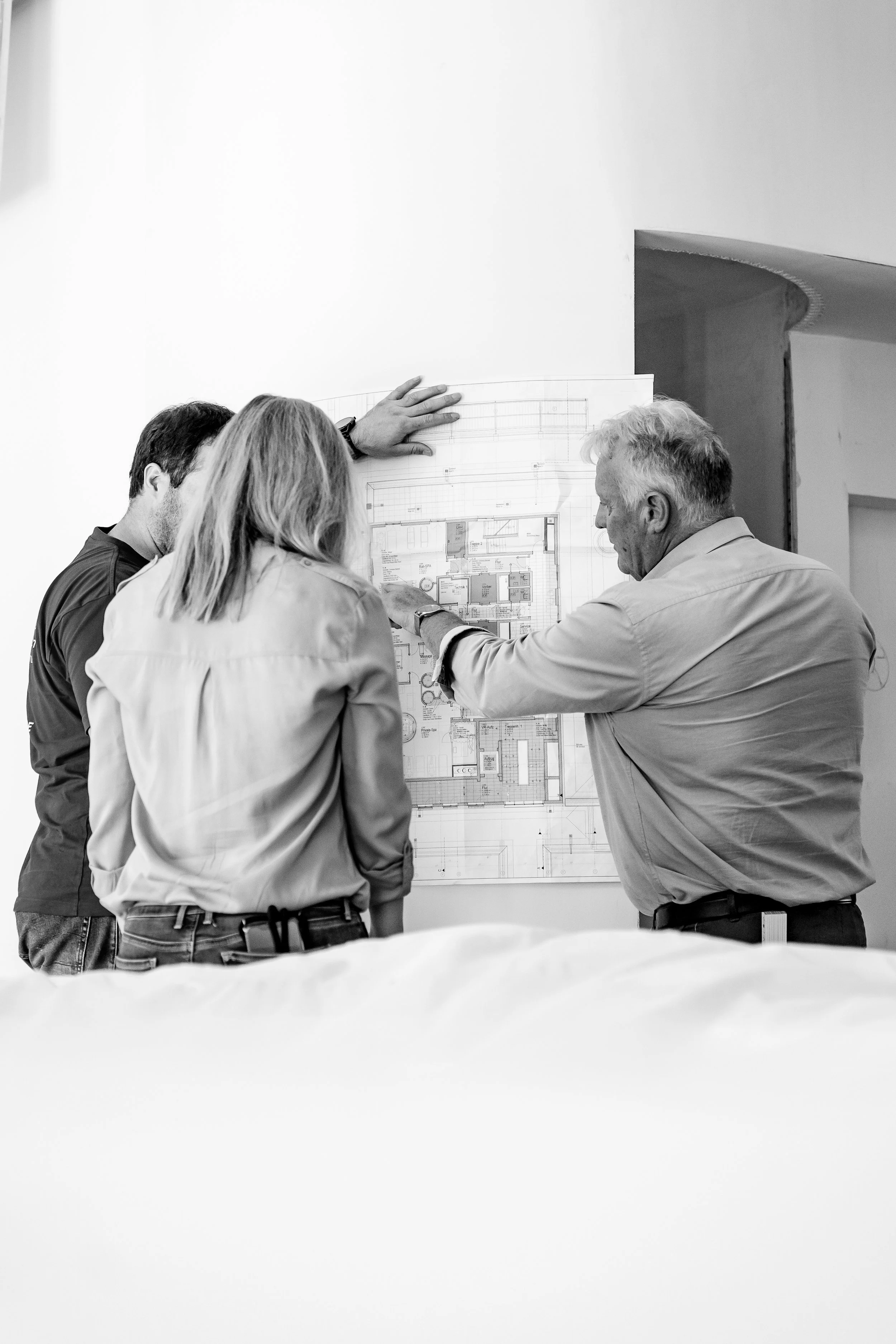 Drei Menschen betrachten einen Bauplan an einer Wand, mit einem älteren Mann, der den Plan erklärt.