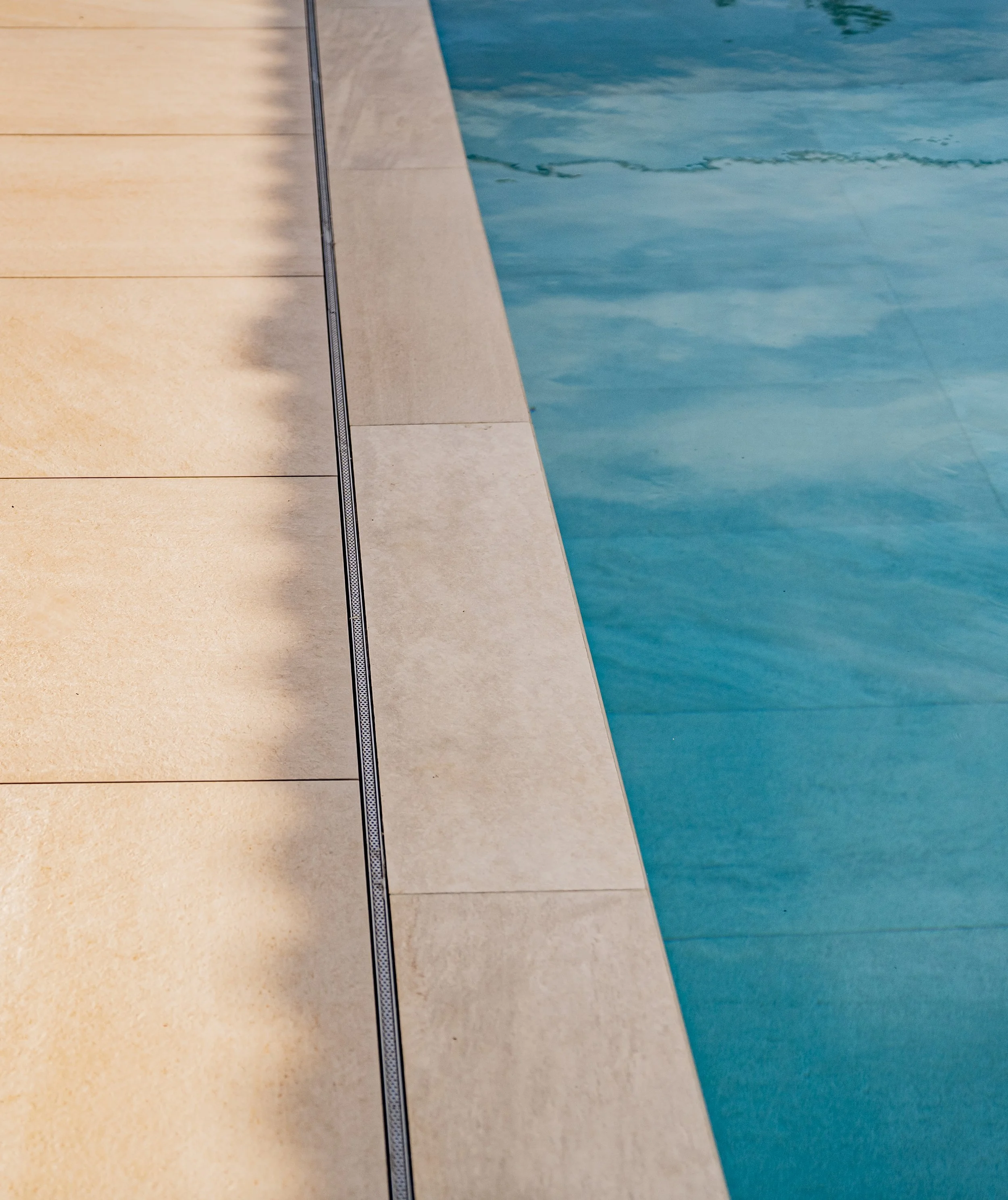 Blick auf den Rand eines Schwimmbeckens, das Wasser ist blau und ruhig, die Beckenumrandung ist beige und glatt.