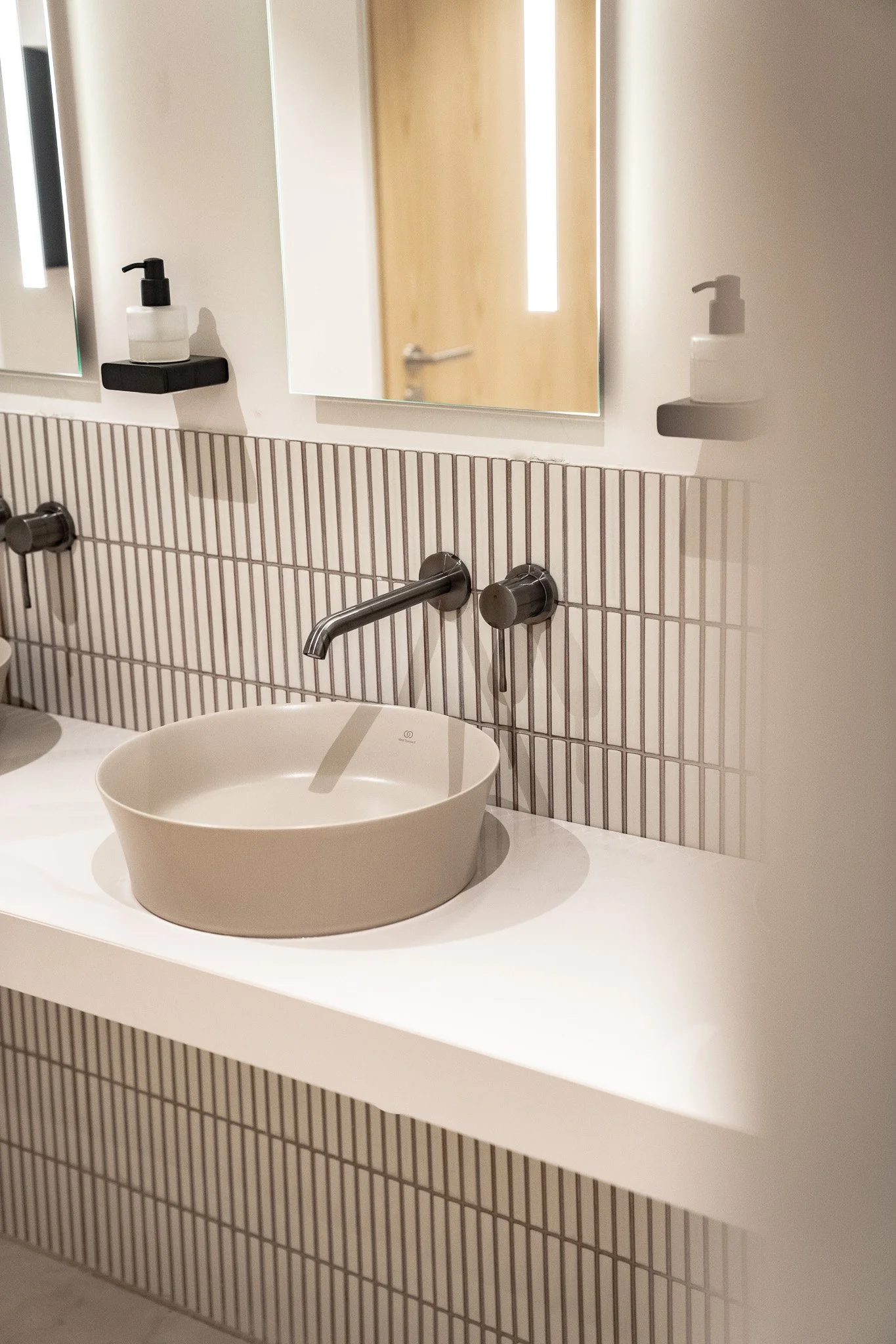 Ein moderiner Waschbeckenbereich mit einer ovalen, beige Waschschale, einem Wasserhahn und zwei Wasserhähnen, einer Wand mit vertikalen schwarzen Linien, zwei Spiegel und Seifenspender auf schwarzen Wandhaltern.