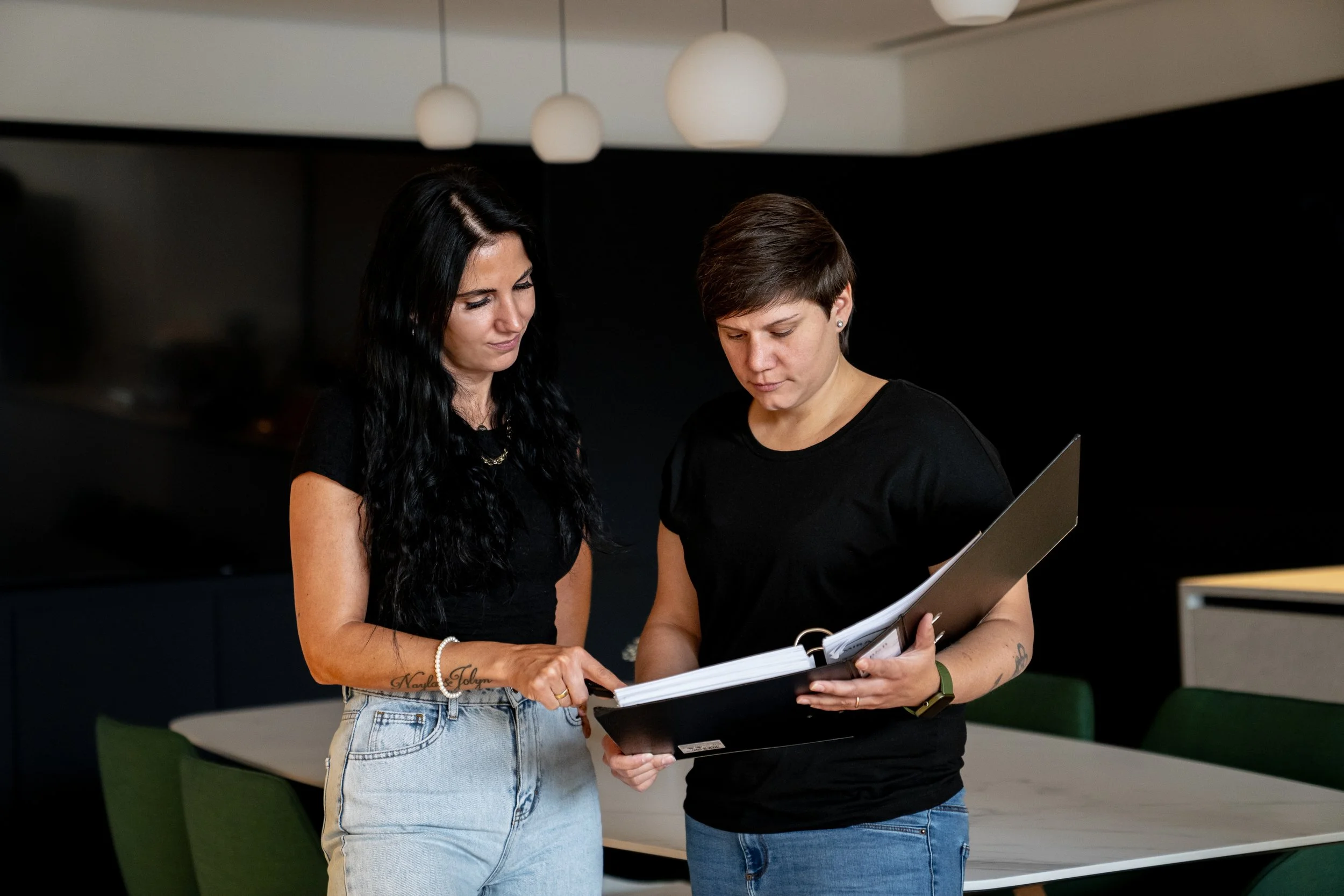 Zwei Frauen, die in einem modernen Raum, vermutlich einem Büro, zusammen einen Aktenordner mit Dokumenten anschauen und besprechen.