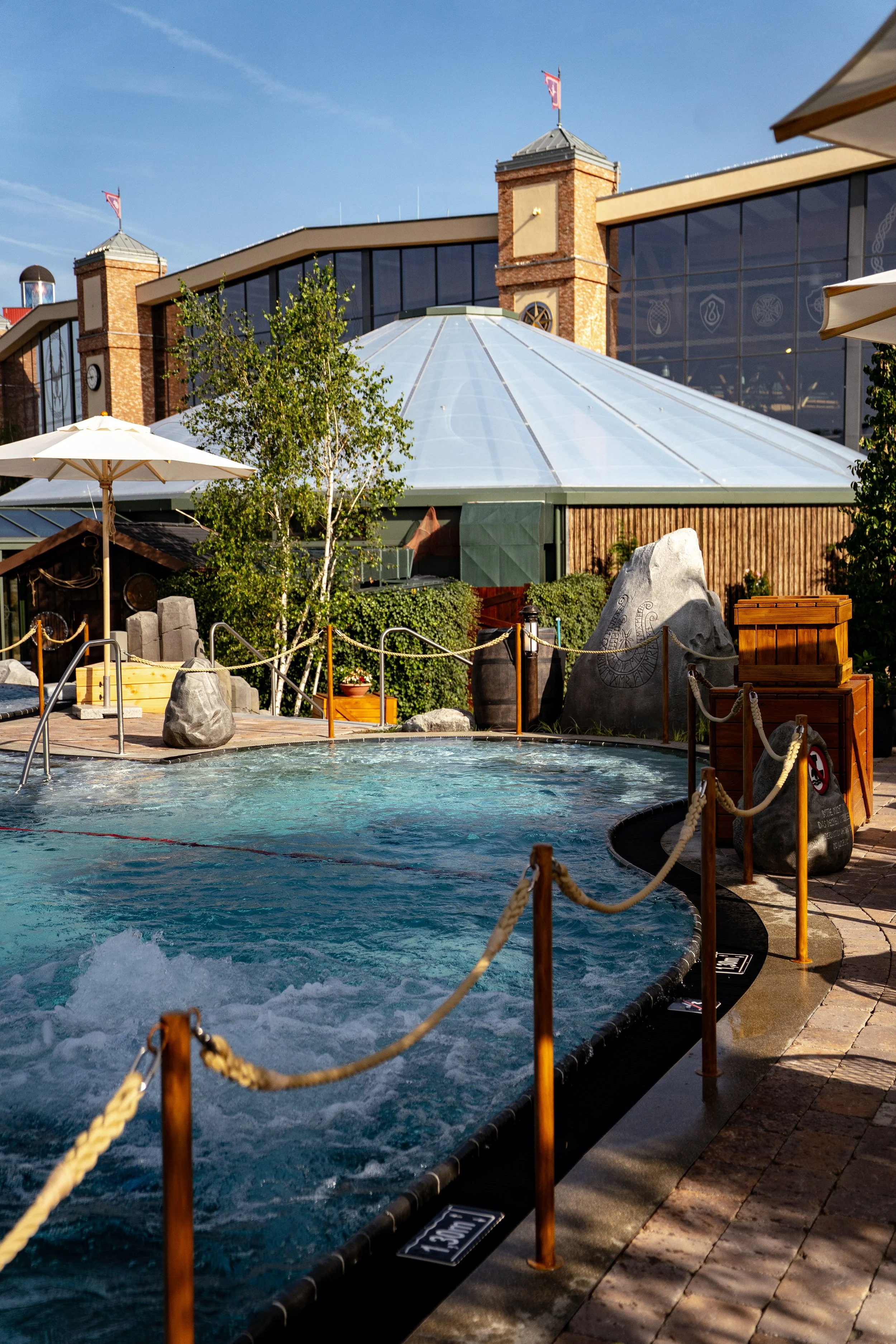 Ein Außenpool mit Wasser, umgeben von Holzpfosten und Seilen, im Hintergrund ein Gebäude mit großen Fenstern und einer gläsernen Kuppel, darunter Bäume und Liegeplätze, sonniges Wetter.