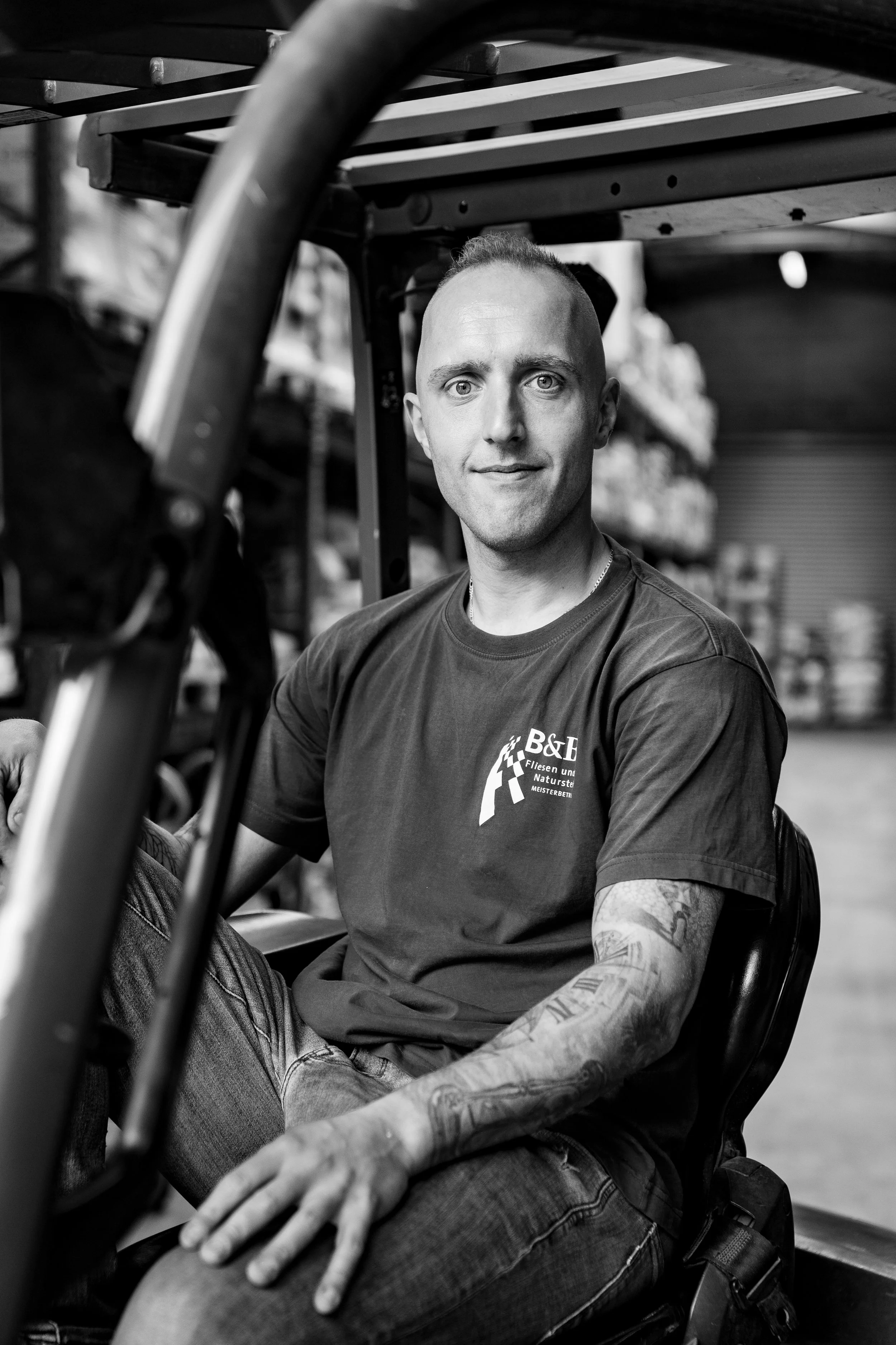 Schwarz-weiß-Foto eines Mannes, der in einem Lager mit Regalen und Waren sitzt, lächelnd in die Kamera schaut.