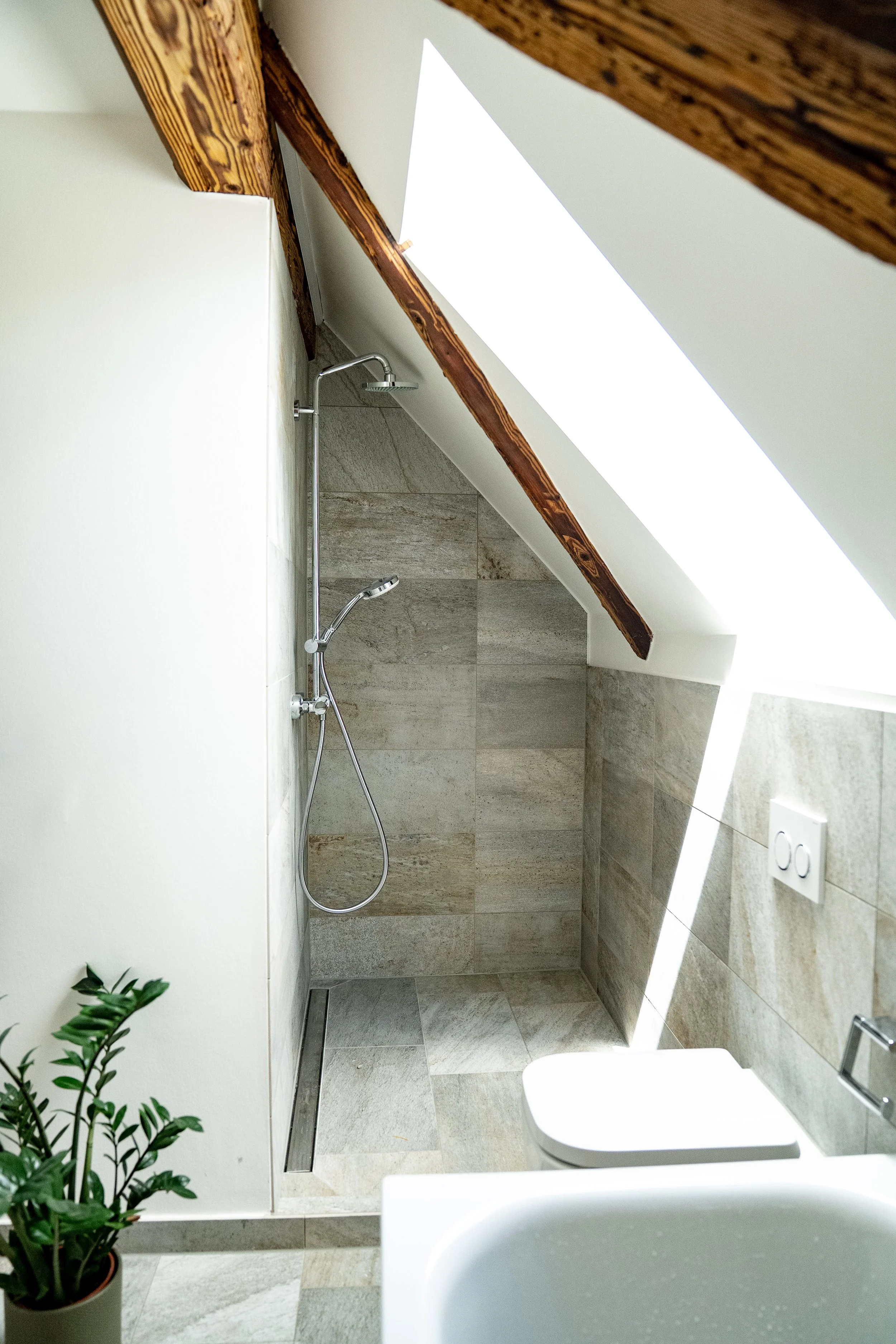 Ein kleines Badezimmer mit einer offener Dusche im Eck, Wandfliesen in Natursteinoptik, eine weiße Wand, Holzdecke und ein Dachfenster. Im Vordergrund ein Waschbecken und eine grüne Zimmerpflanze.
