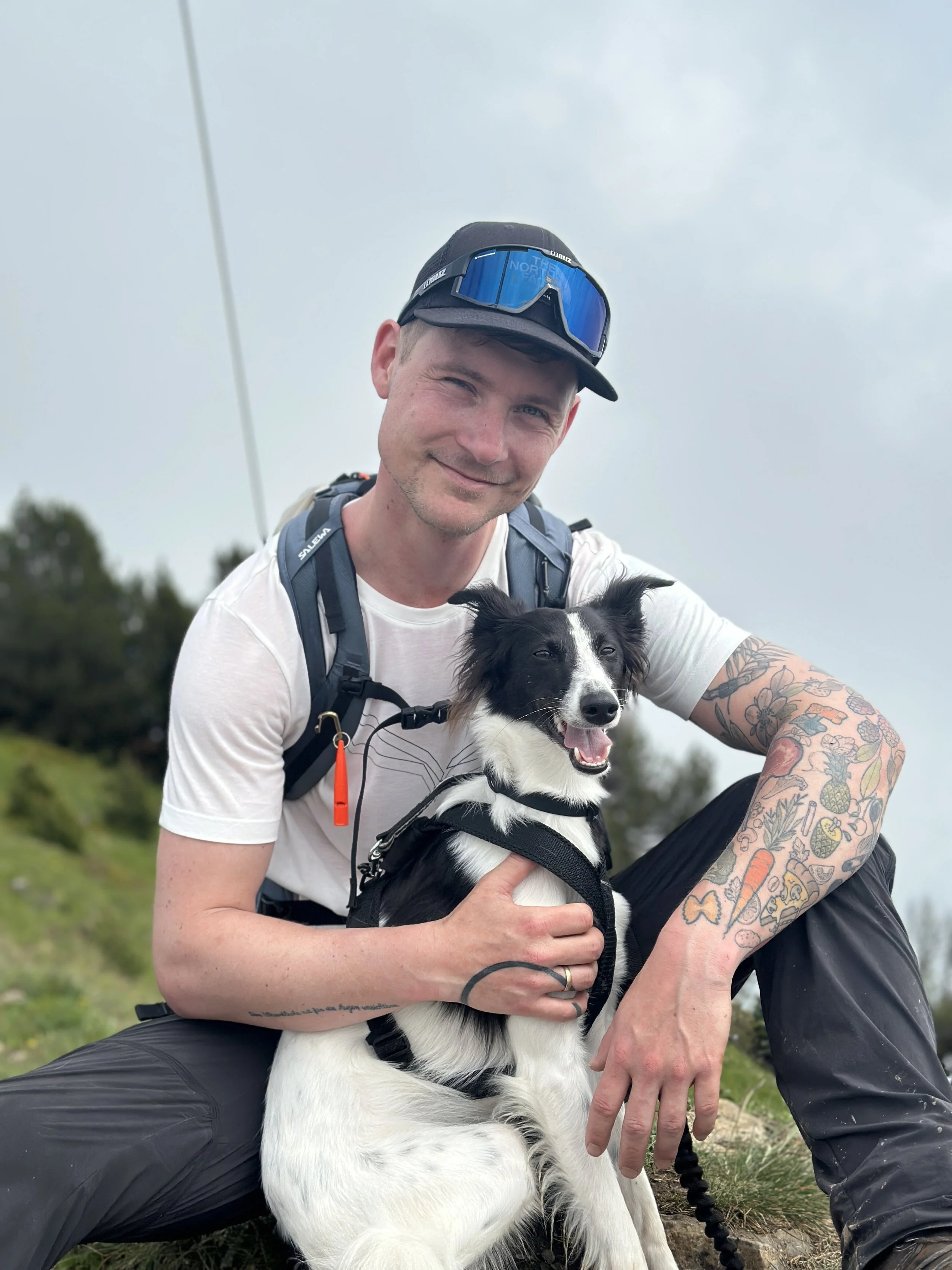 Ein Mann in Outdoor-Kleidung und mit Tattoos, der auf einer grünen Wiese sitzt und einen schwarz-weißen Hund hält, beides lächelnd bei bewölktem Himmel.