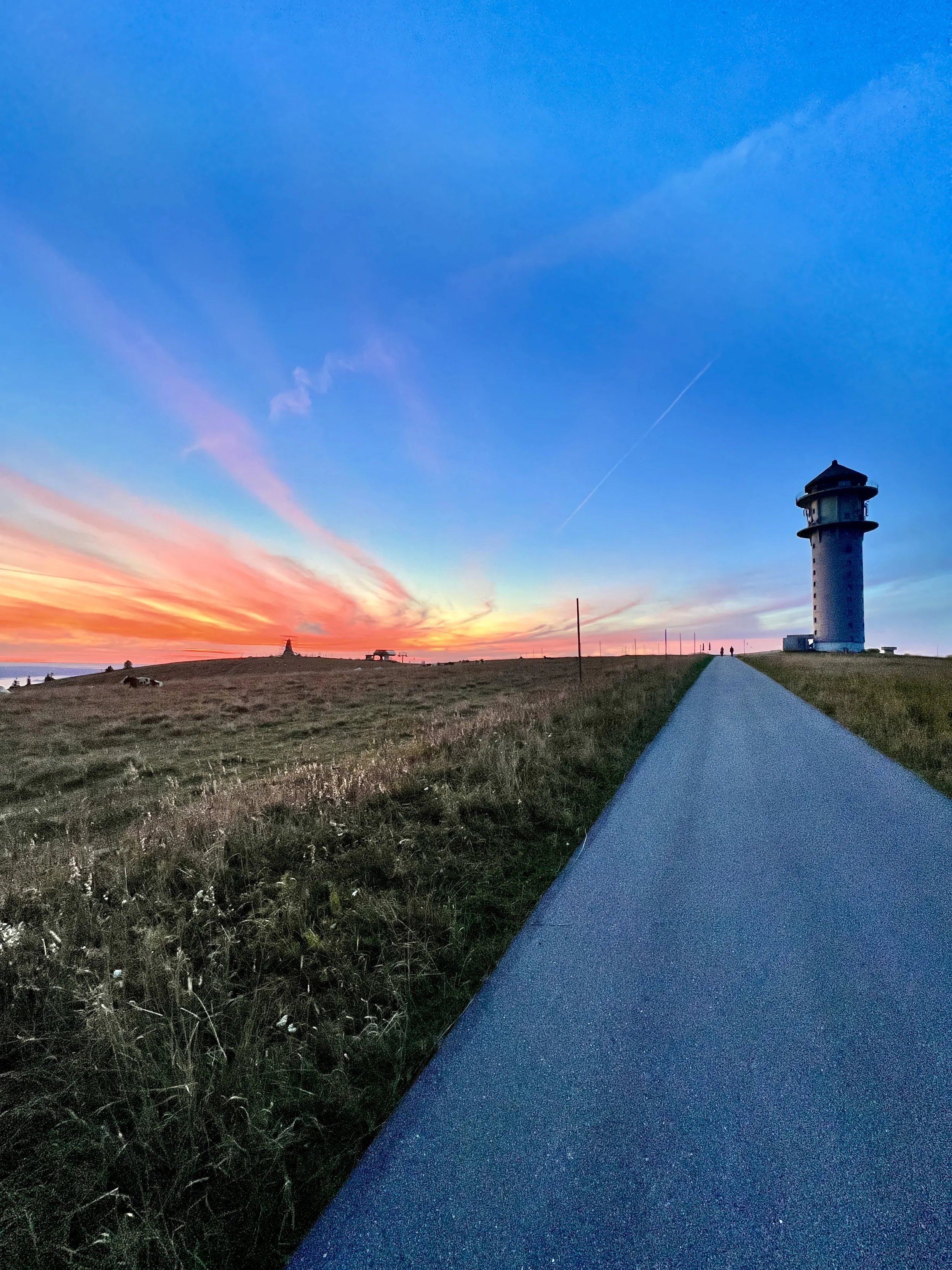 Blick auf einen Leuchtturm bei Sonnenuntergang mit buntem Himmel und einer mit Asphalt bedeckten Straße, die Richtung Leuchtturm führt.