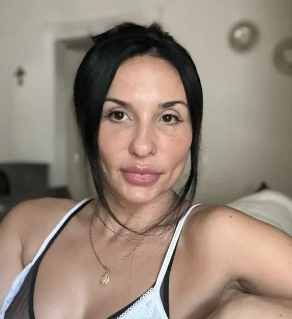 A woman with dark hair and light skin taking a selfie in a room with a bed in the background.