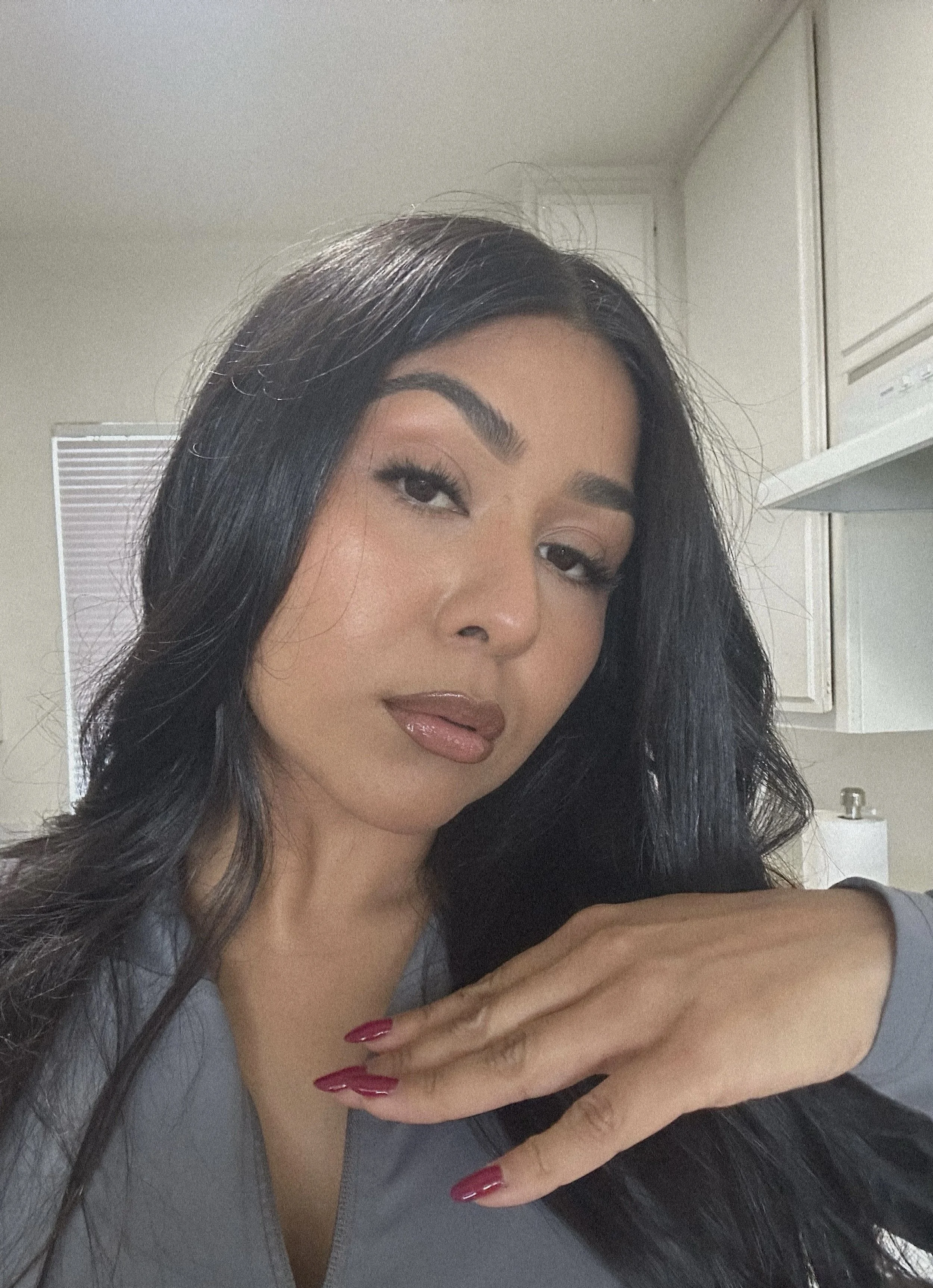 A woman with long black hair, wearing makeup, and red nail polish, resting her hand near her face, in a room with white cabinets and a window with blinds in the background.