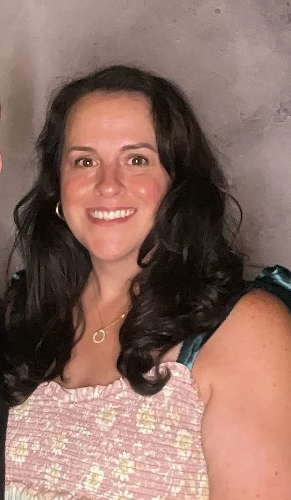 A smiling woman with long dark curly hair wearing a pink dress with a floral pattern and black straps, and a gold necklace, posing in front of a plain background.