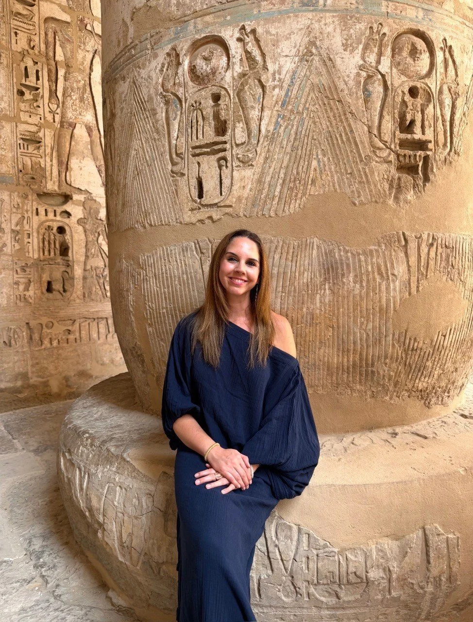 A woman in a navy blue dress sitting on a stone bench in front of an ancient Egyptian archaeological site with hieroglyphics and carvings on the walls and a large round stone structure.