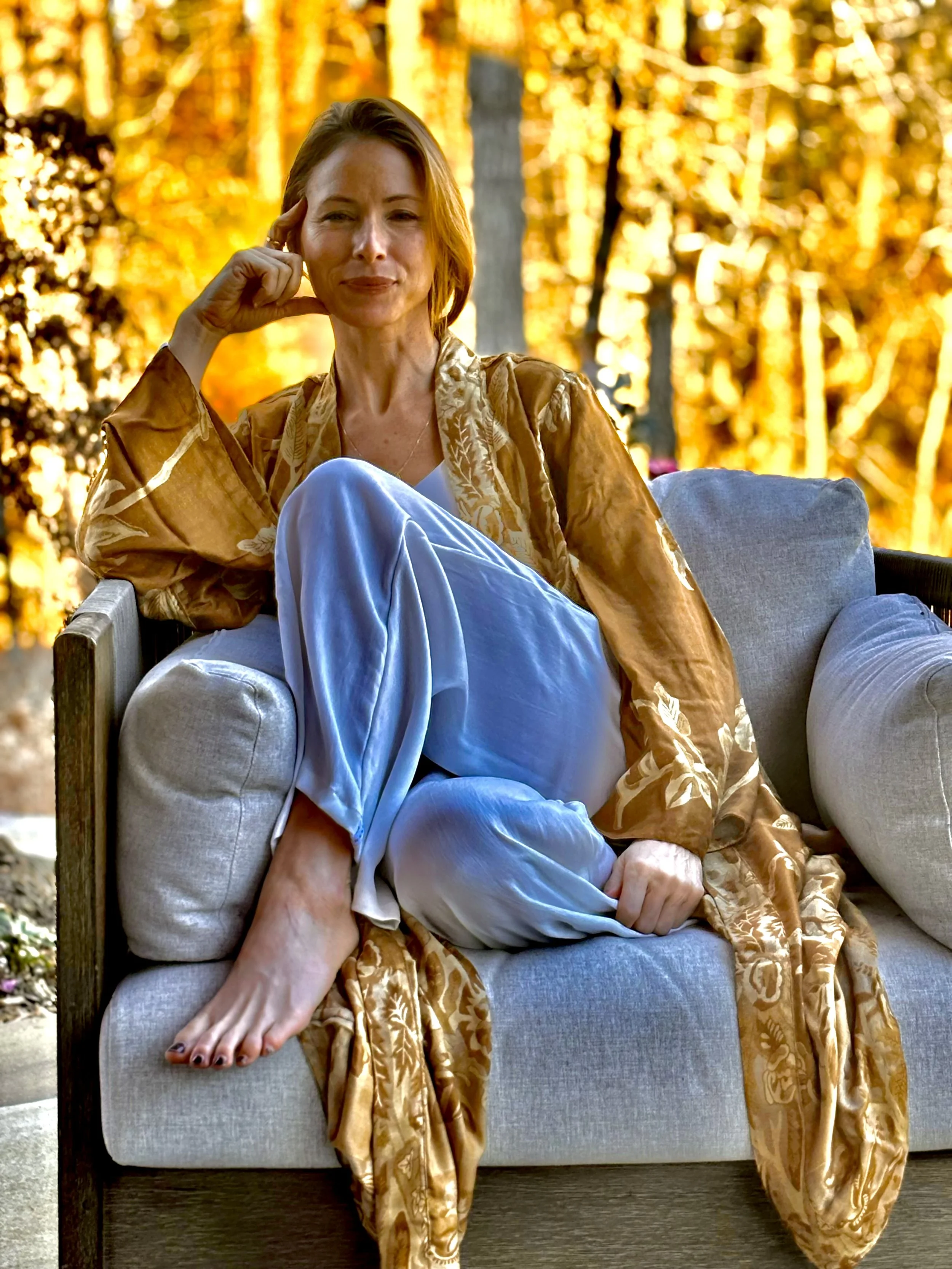 A woman with short hair sitting on a sofa outdoors during autumn, wearing a brown patterned robe over light blue pajamas, with her right leg crossed over her left, and her right hand resting near her temple.