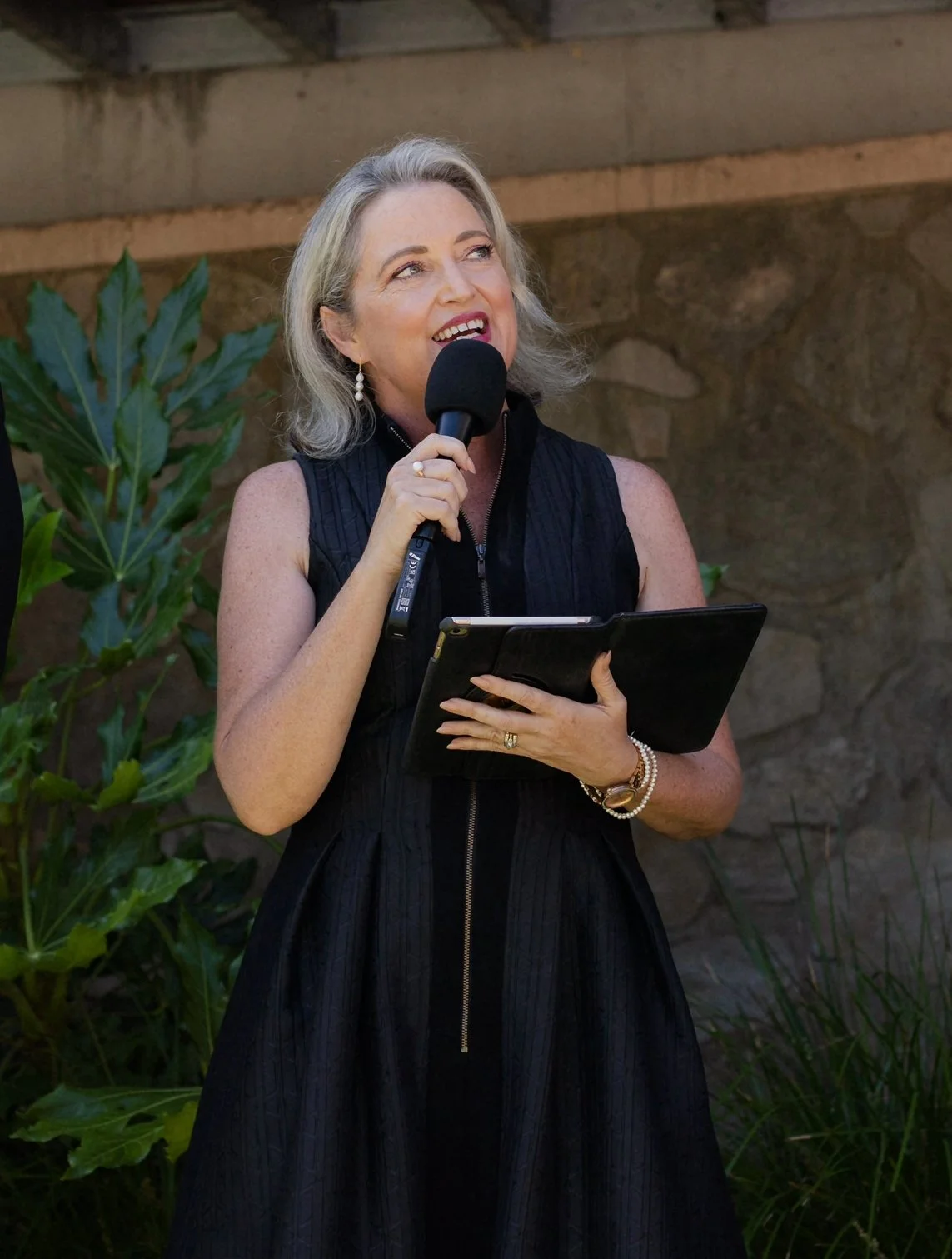 A woman with blonde hair smiling at an indoor event, wearing a black dress and pearl earrings.