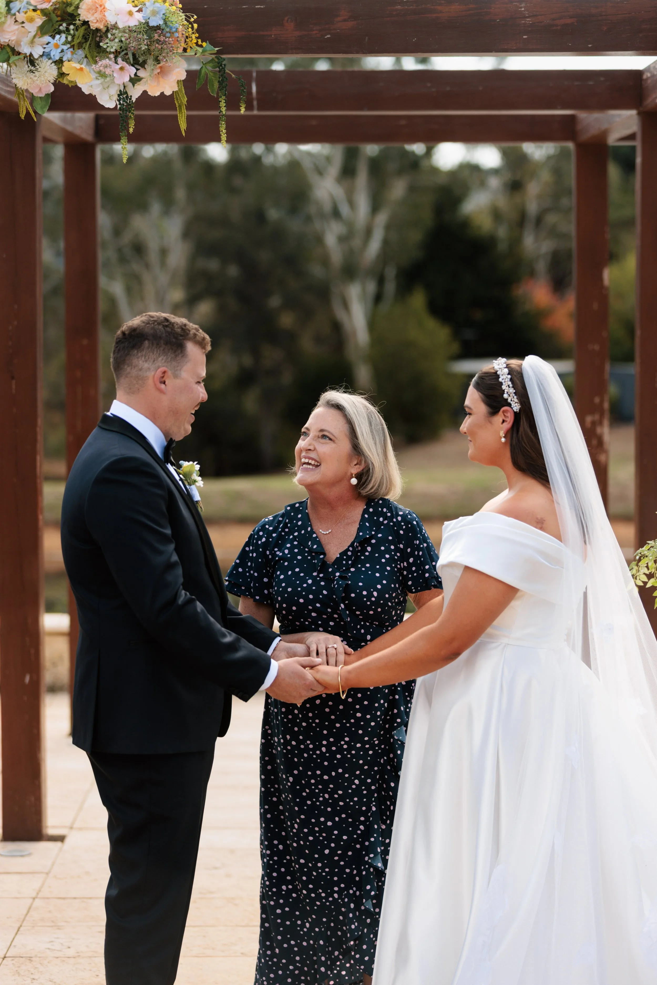 A wedding celebrant holding hands with a couple underneath an arbour
