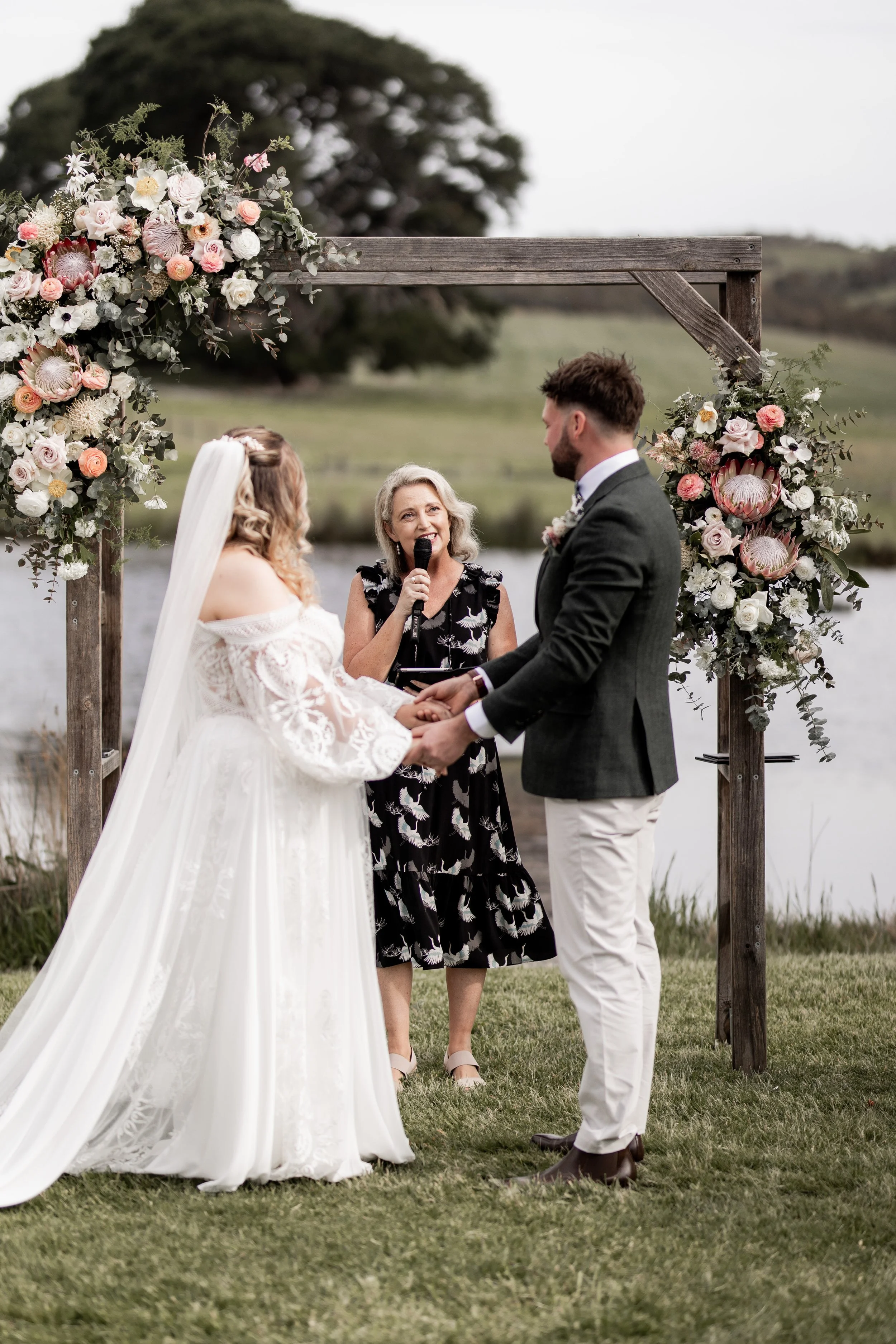 A couple getting married outdoors by a lake, with a woman officiant holding a microphone, standing under a wooden wedding arch decorated with pink and white flowers.