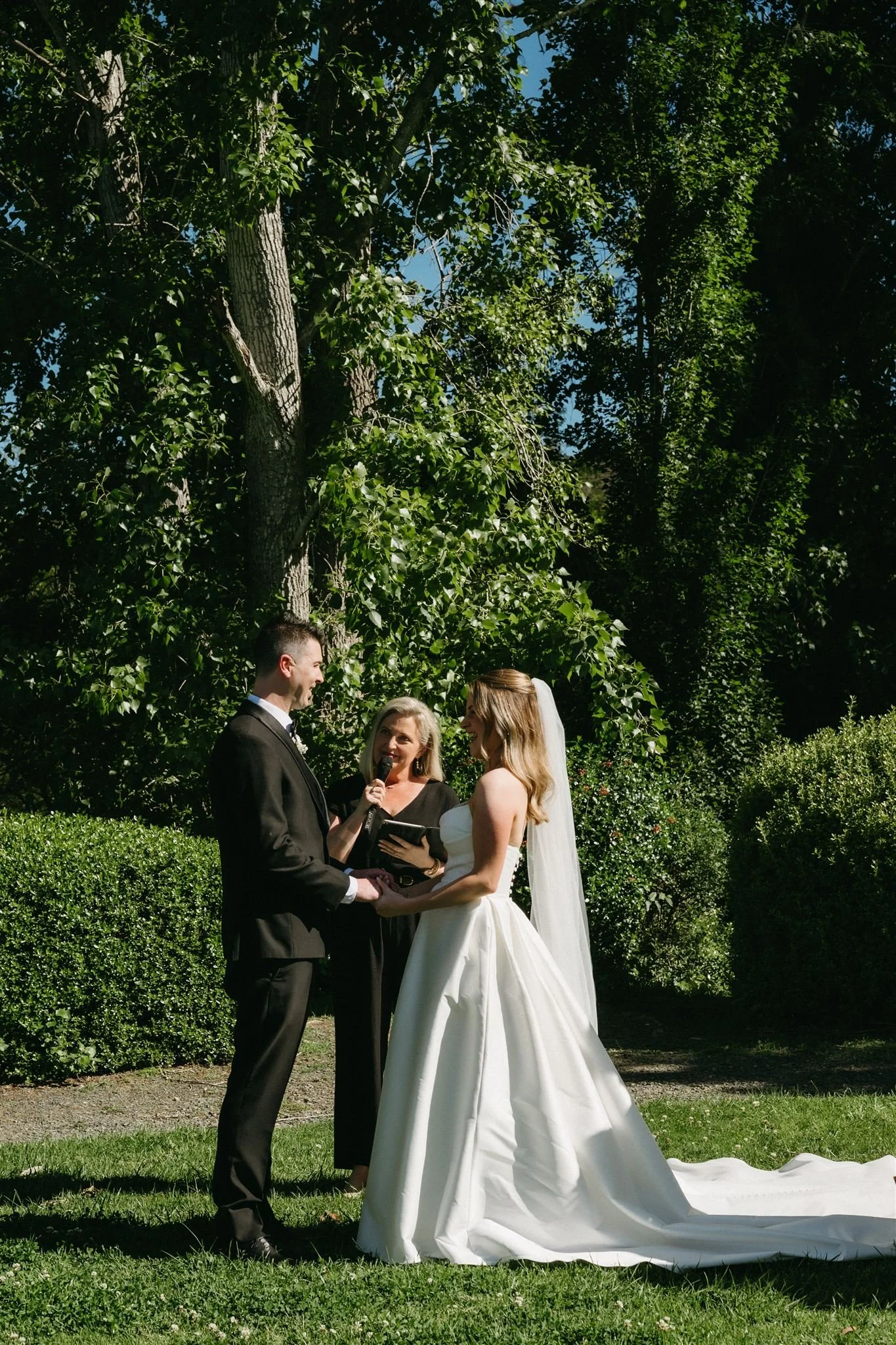 A newlywed couple in wedding attire holding a black poodle dog outdoors. The groom is wearing a black tuxedo with a bow tie, and the bride is wearing a white wedding dress with ruffled sleeves and a veil. The poodle is dressed in a white outfit with bows in its ears.