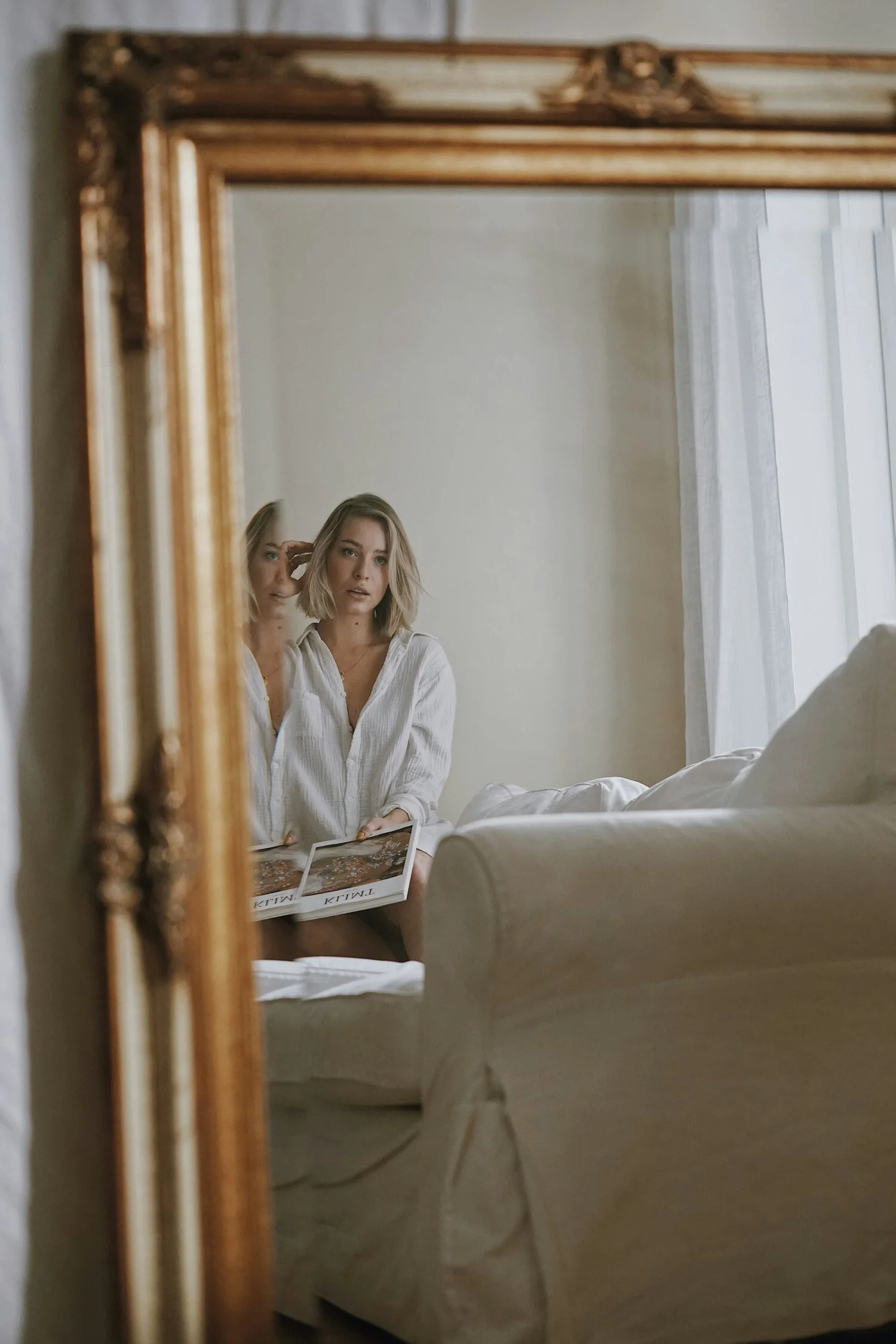 A woman with short blonde hair in a white shirt is sitting on a white sofa, looking into a mirror, holding a magazine, in a bright room with a large window and white curtains.