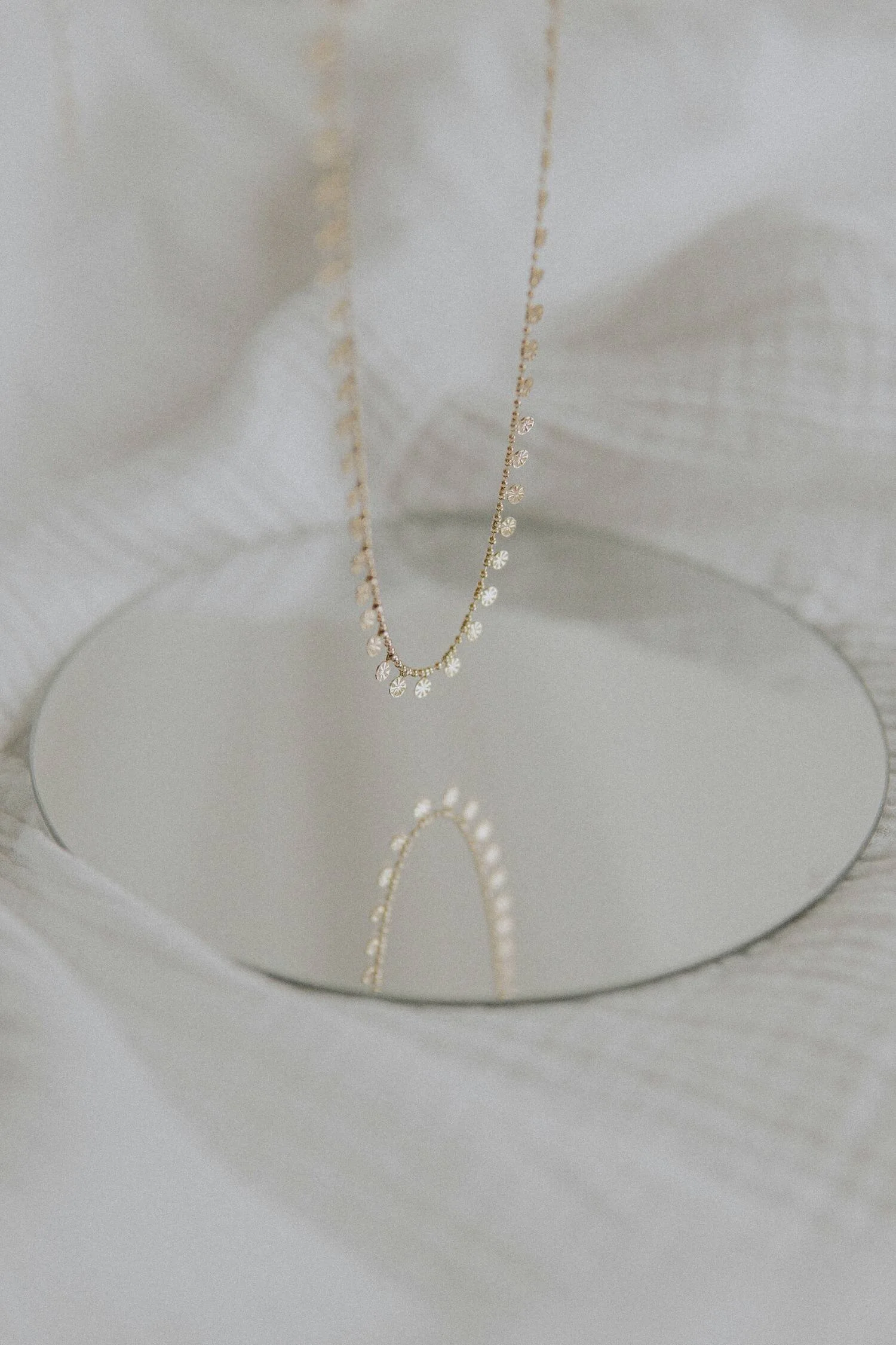 A delicate gold necklace with small round diamonds hangs over a round mirror, reflecting the necklace's pattern.