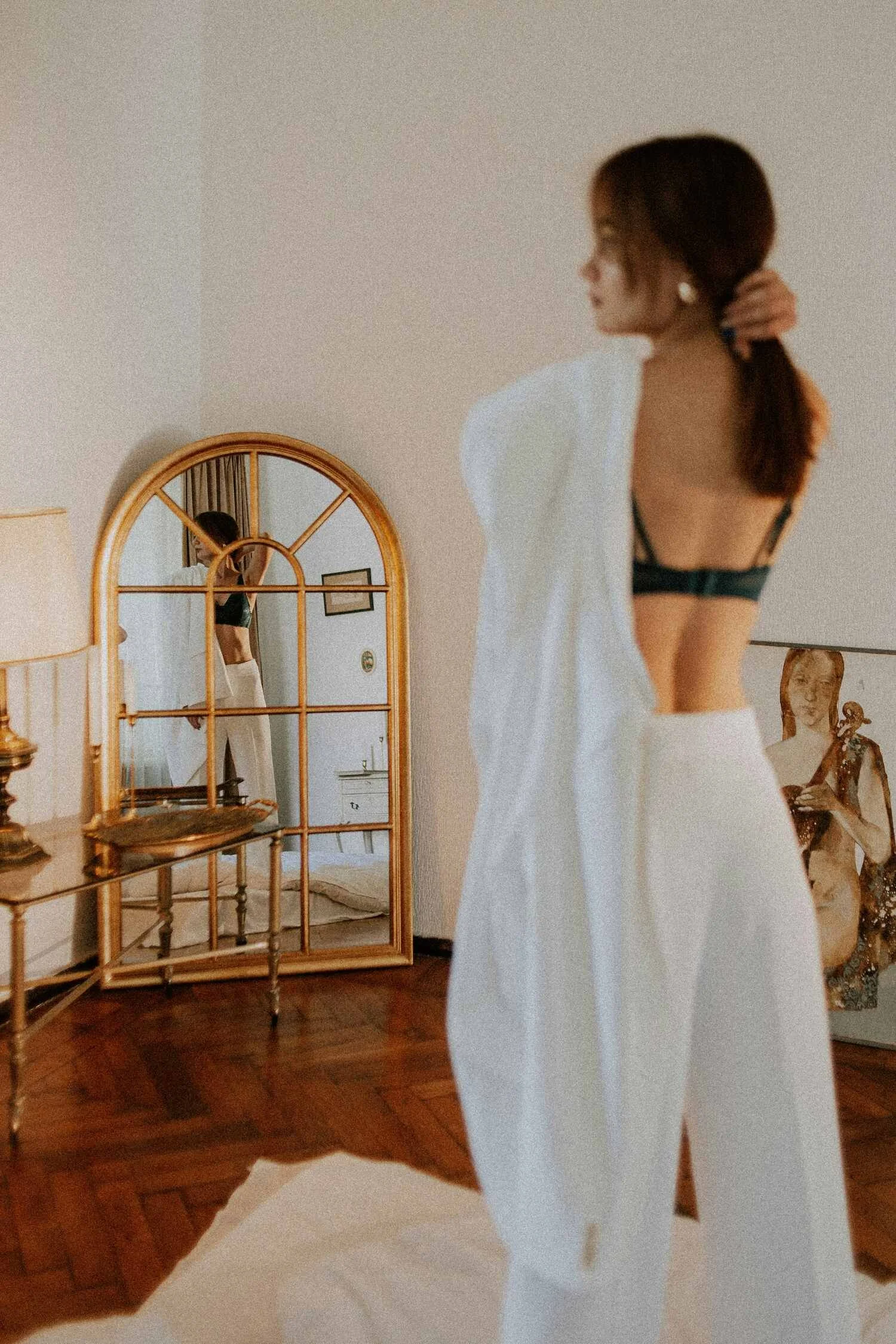 A woman in black bra and white pants adjusting her hair in front of a mirror, with her reflection visible in the mirror, in a room with wooden floor and beige walls.