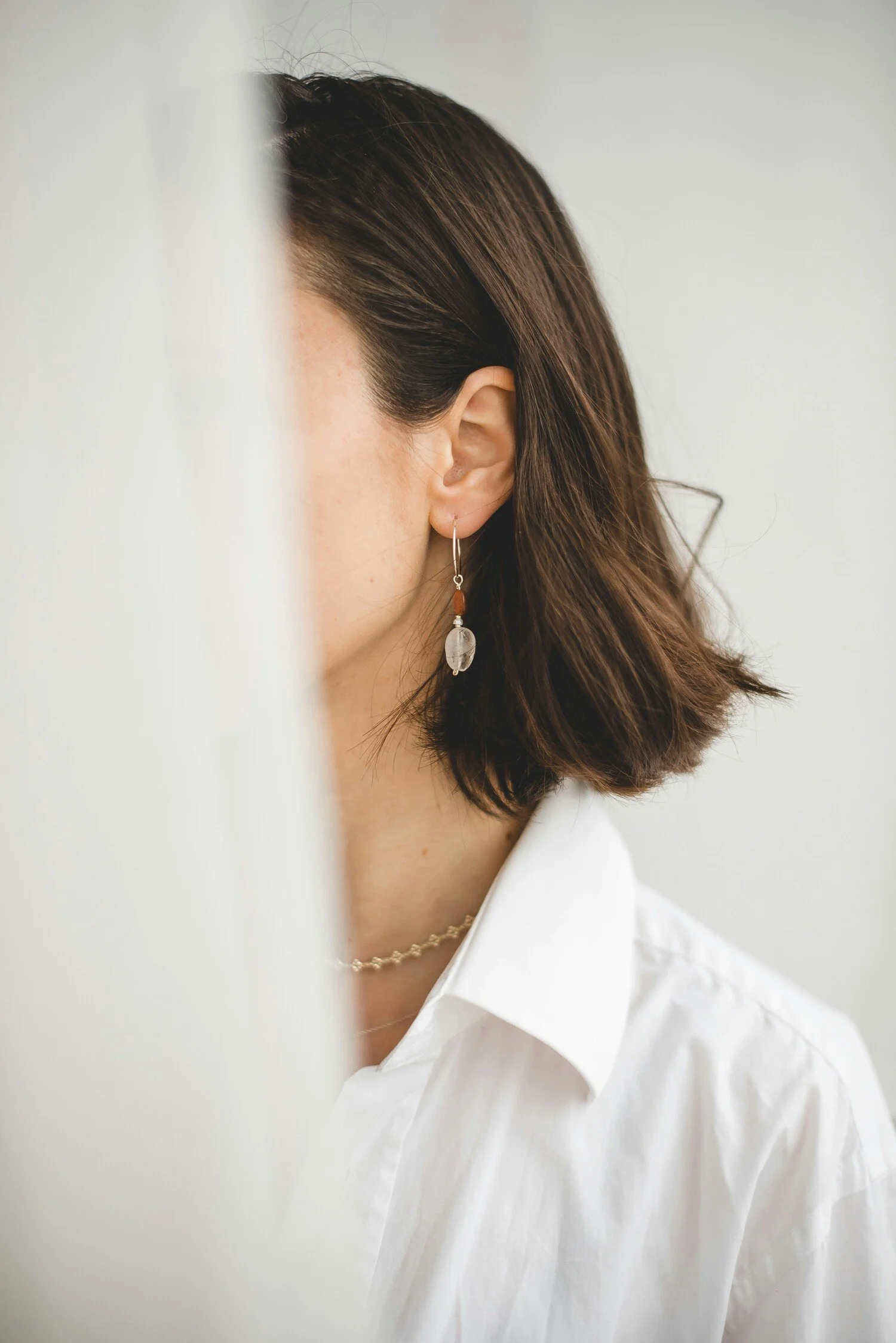 Close-up of a woman's face partly hidden behind a white curtain, showing her ear with an earring and her brown hair, white shirt, and a necklace.