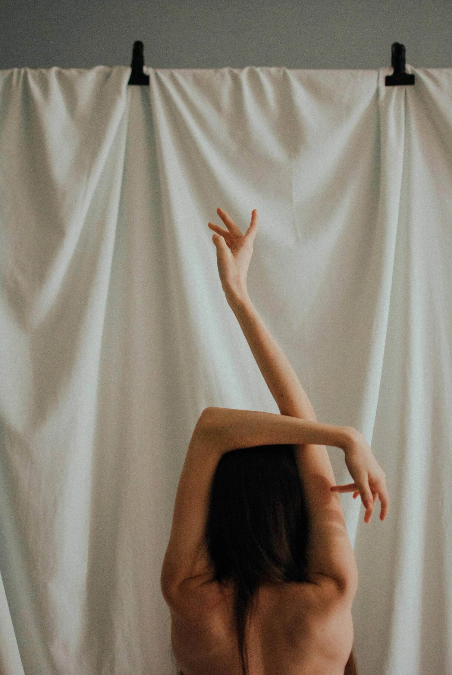 A person with long dark hair and bare shoulders standing in front of a white fabric backdrop, with one arm raised and fingers extended gracefully.
