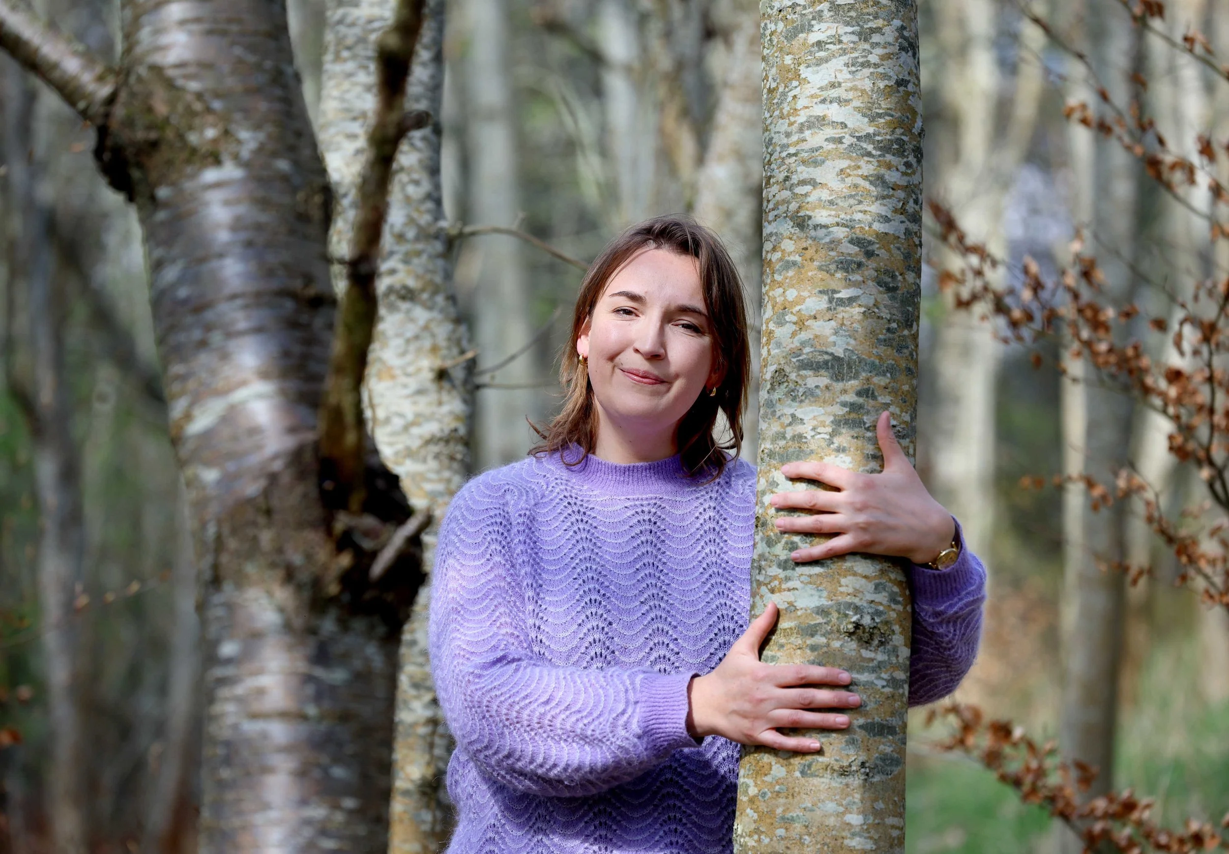 Close-up of a woman with brown hair and brown eyes, wearing a colorful, patterned sweater, outdoors among trees and greenery.