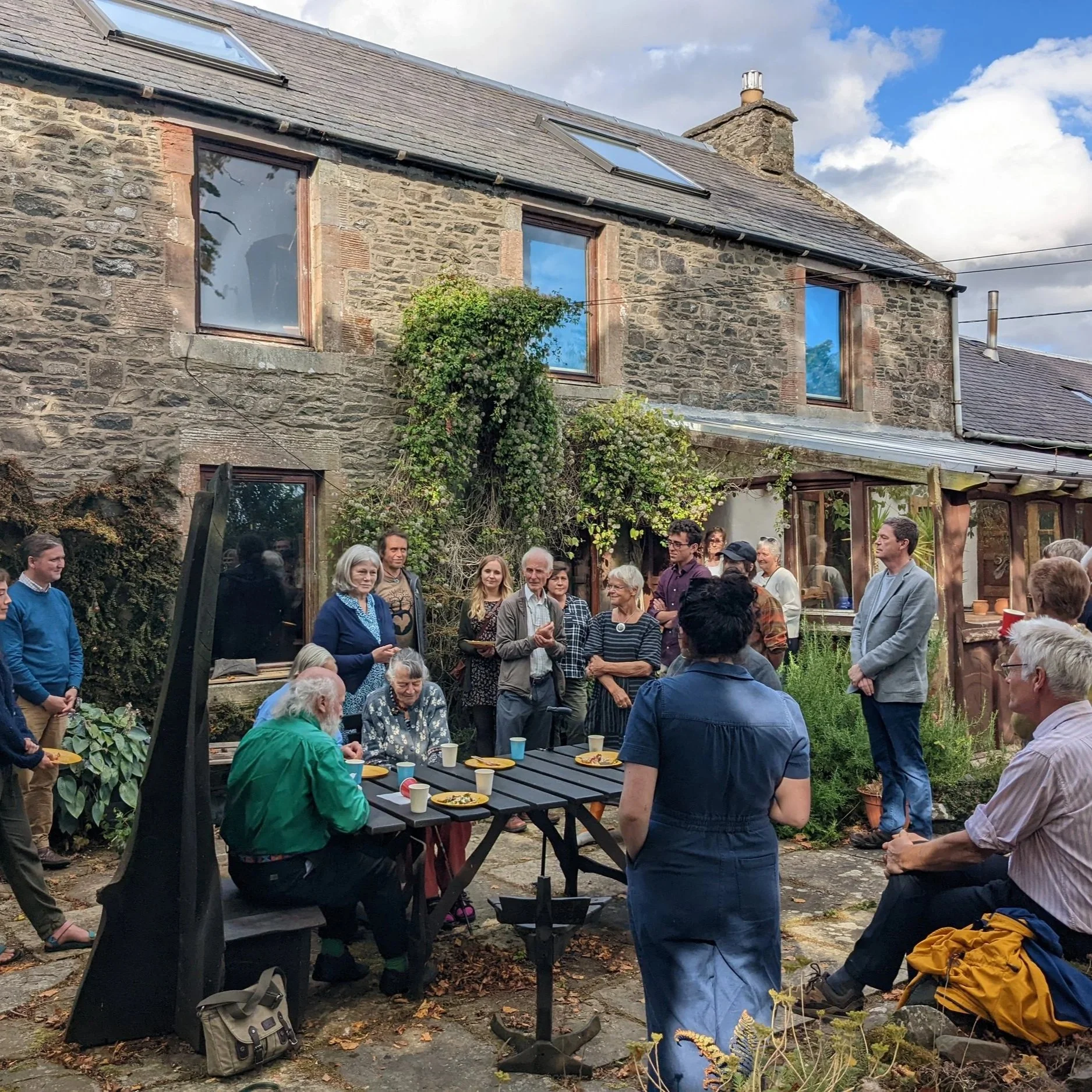 Group of people gathered outdoors in a courtyard for a social event, with some seated at a table and others standing, in front of a stone house with large windows and a garden.