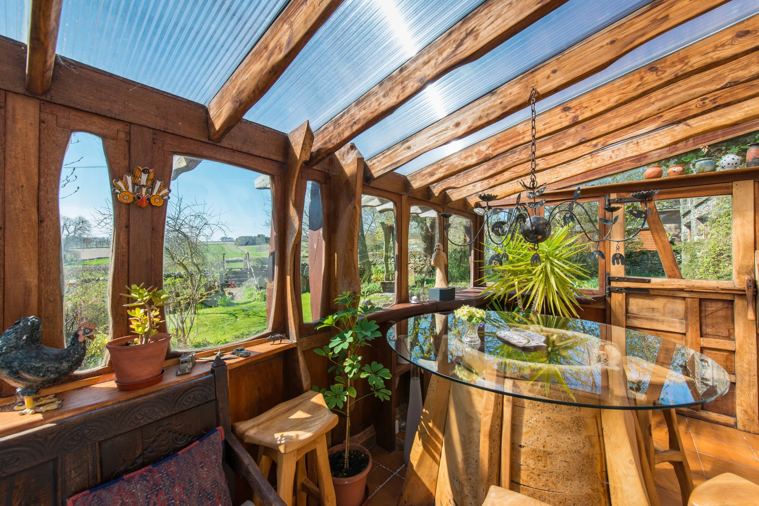 Interior of a sunny wooden greenhouse with a glass table, potted plants, sculptures, and a chandelier, overlooking a green outdoor landscape.