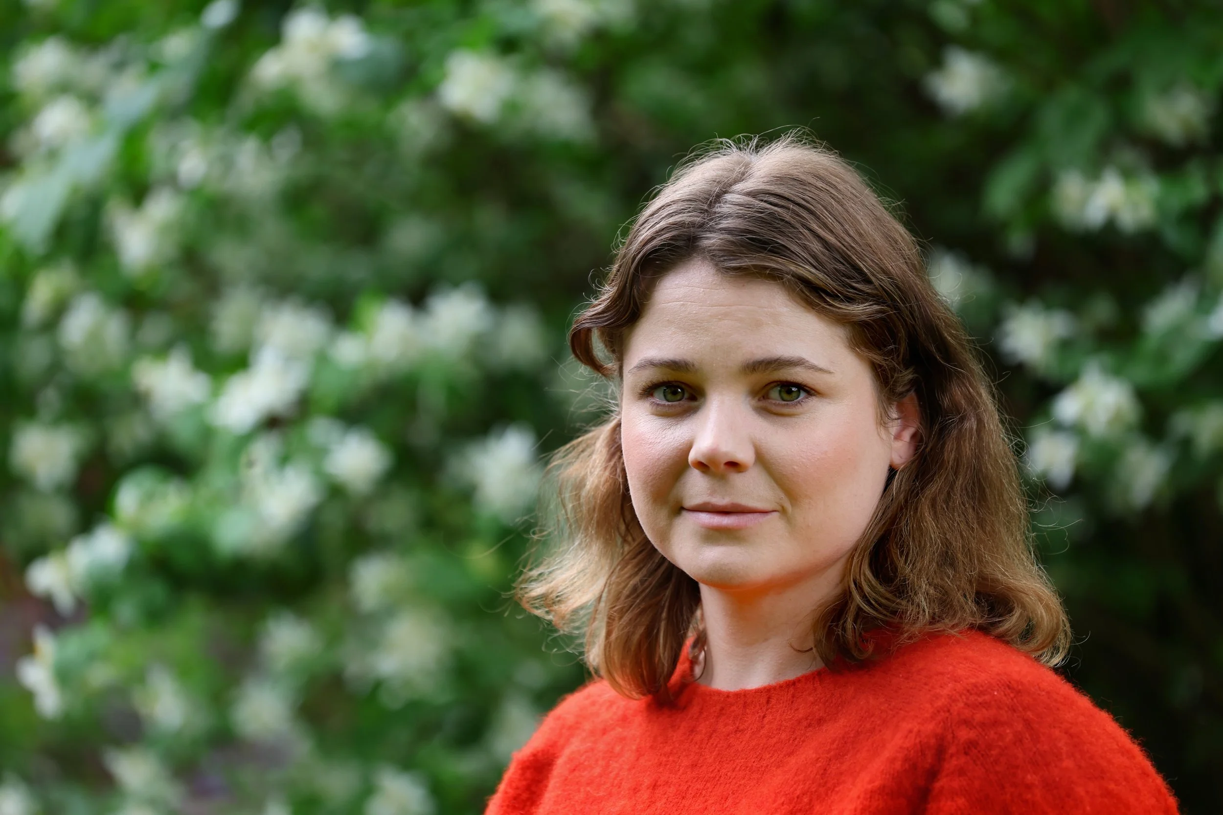 A portrait of a young woman with brown wavy hair and green eyes, wearing a red sweater, standing outdoors with green foliage and white flowers in the background.