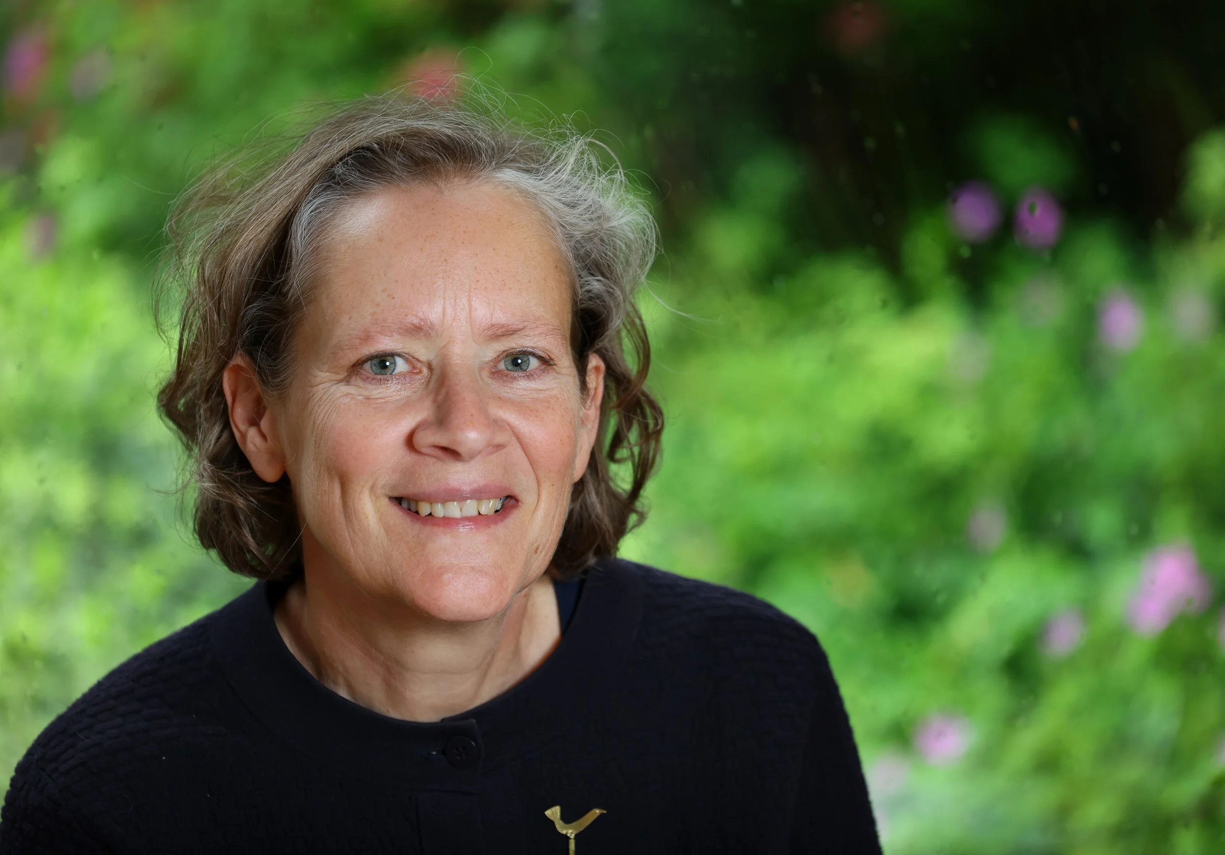 Portrait of a woman with short brown curly hair, smiling, wearing a black top with a gold bird pin, outdoor background with greenery and purple flowers.
